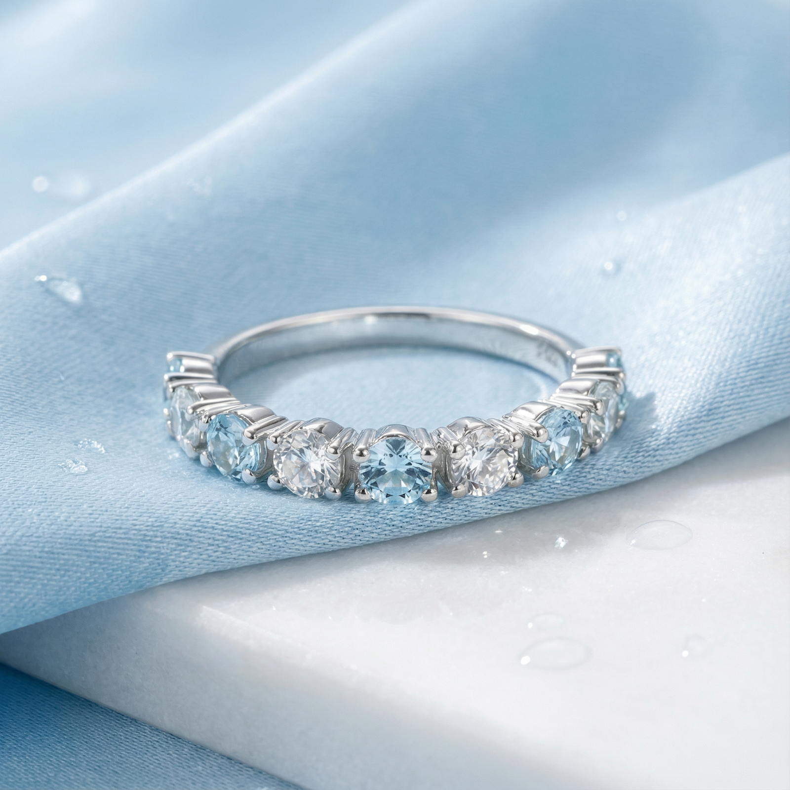 Round cut simulated aquamarine and silver half-eternity band on blue fabric and white marble.