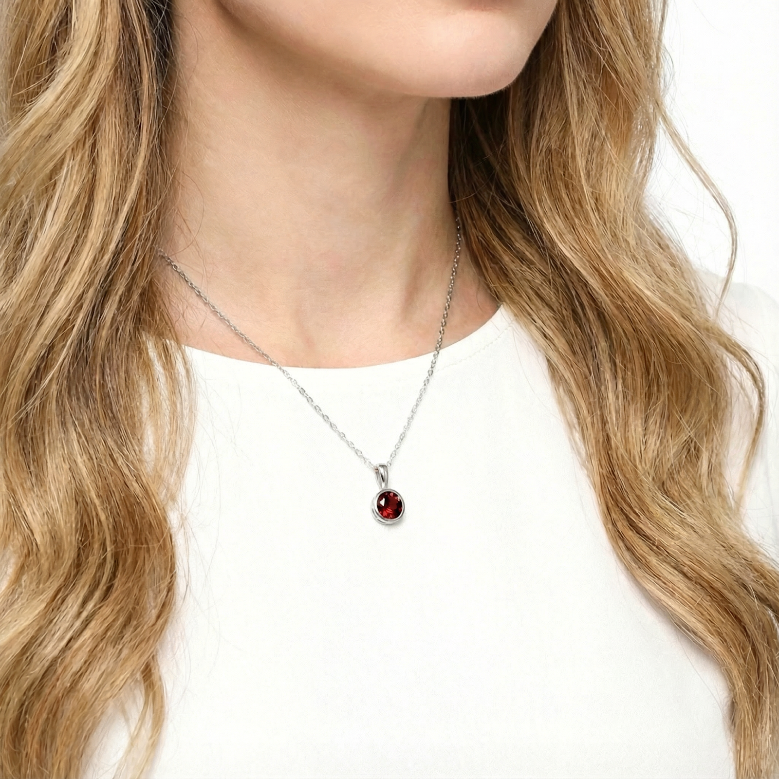 Woman wearing the Alanza round cut red garnet pendant in sterling silver on a delicate chain against a white top.