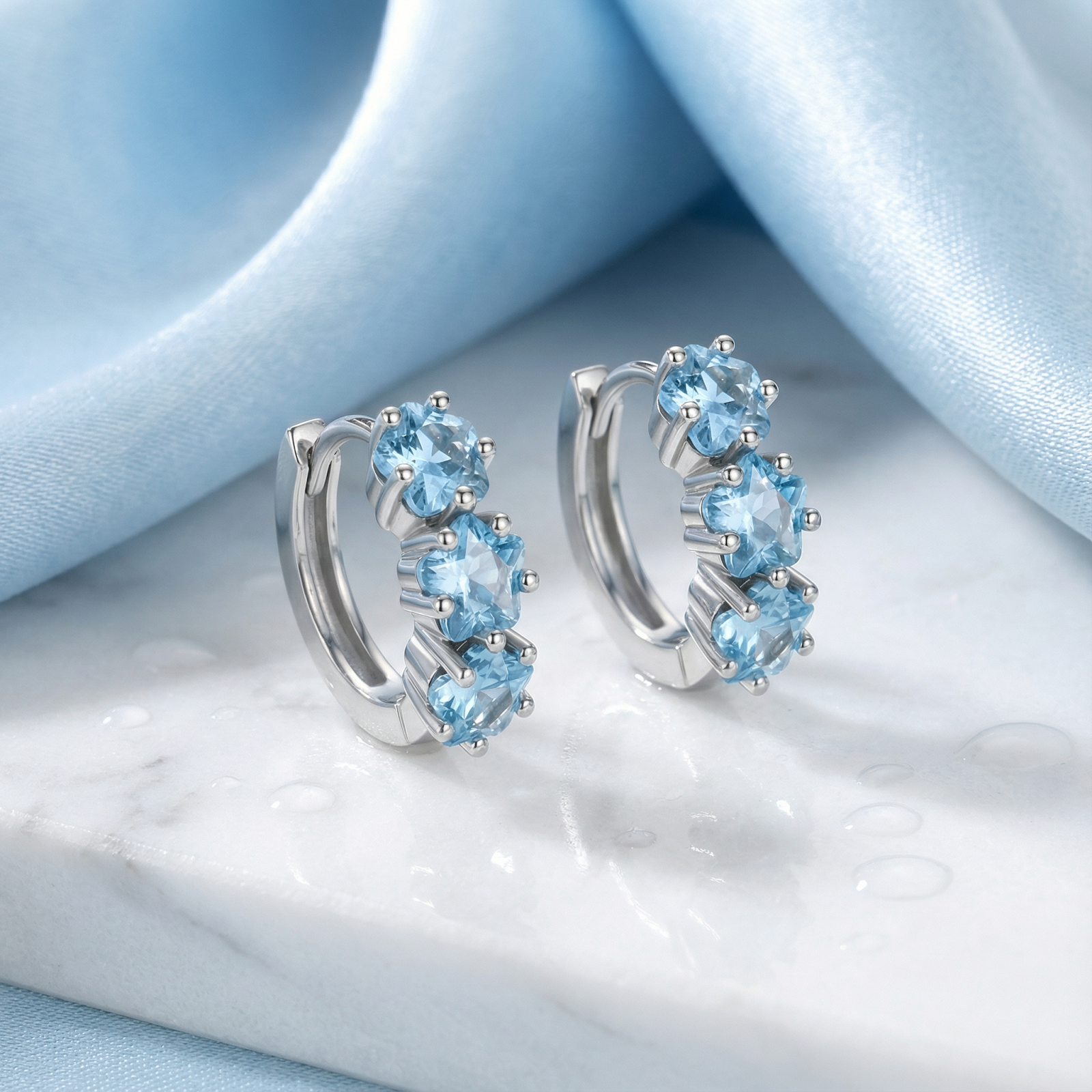 Sterling silver Astra Star Hoops with star-cut light blue aquamarine stones on a marble surface with blue fabric.