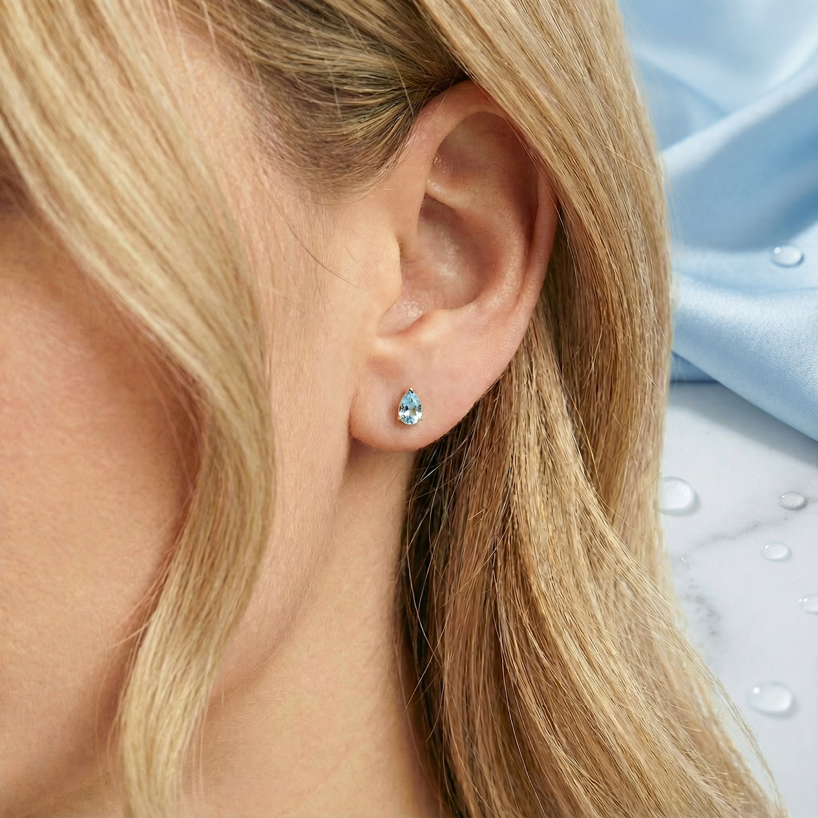 Close-up view of a pear cut simulated aquamarine teardrop earring in 18K gold vermeil worn on an ear.