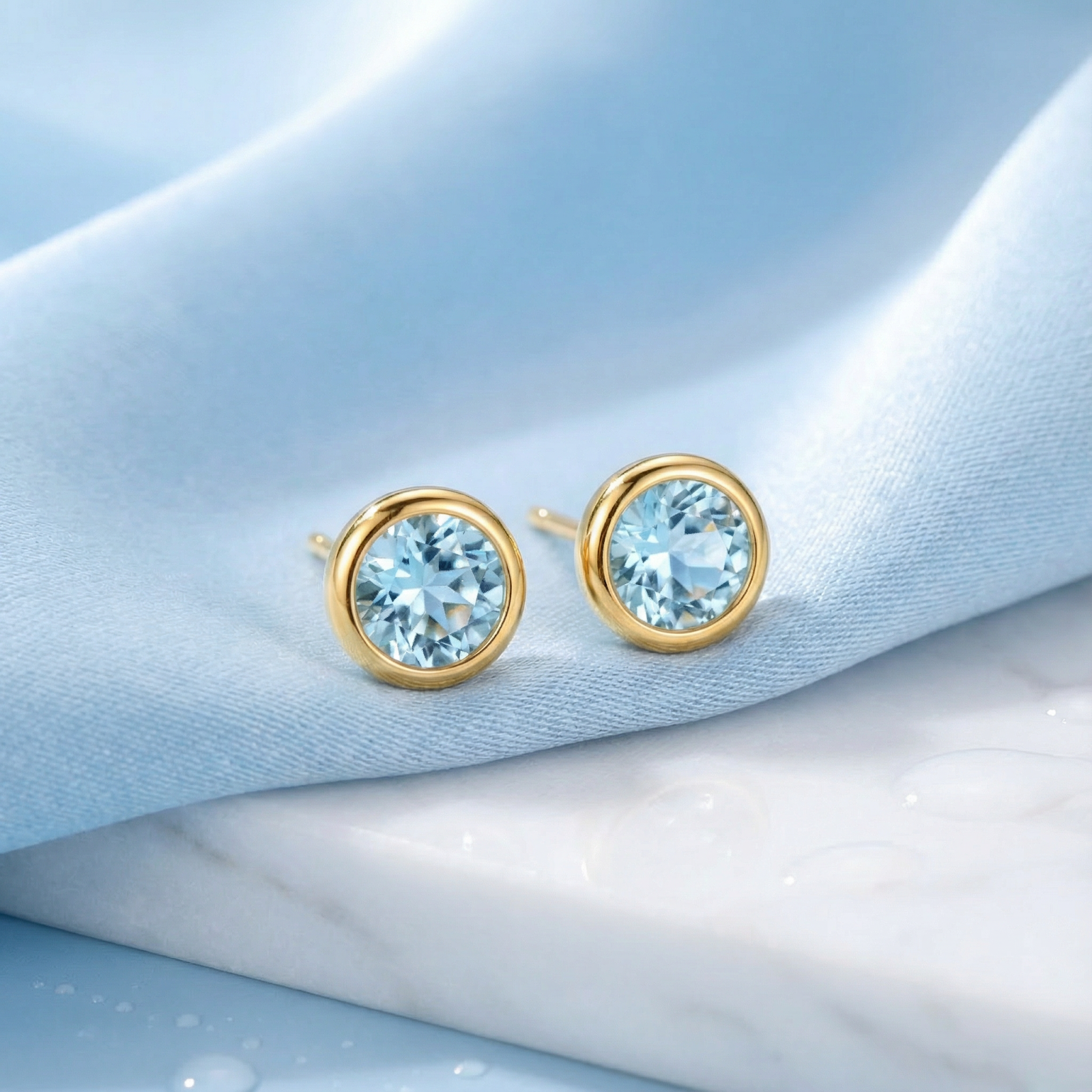 Front view of round simulated aquamarine stud earrings in 18K gold vermeil on a blue silk and marble surface.