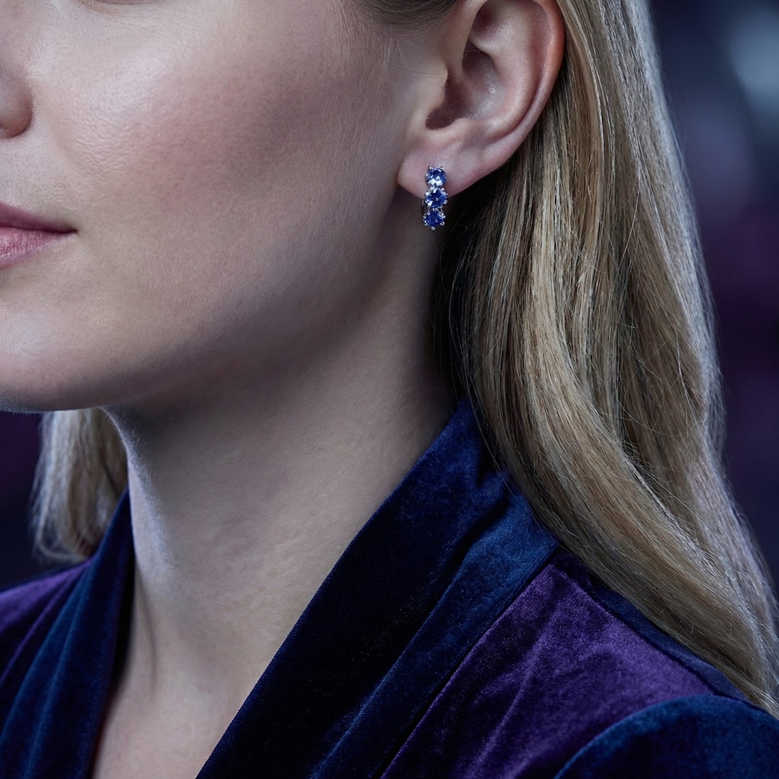 Woman wearing sterling silver star huggie earrings with blue simulated tanzanite stones and a purple velvet top.