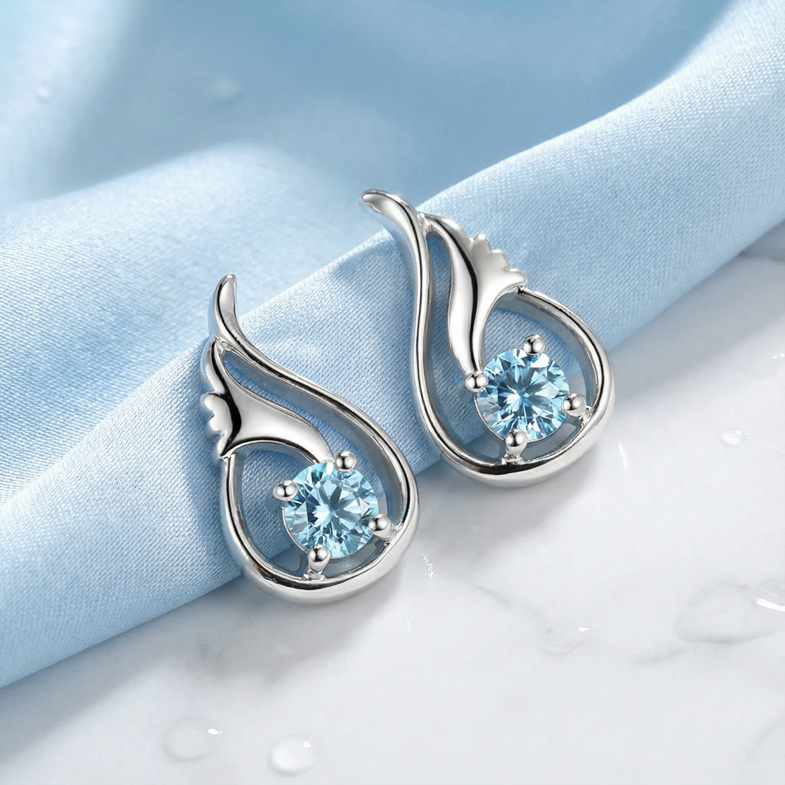 Angled view of sterling silver drop earrings with round-cut light blue simulated aquamarine on blue fabric.
