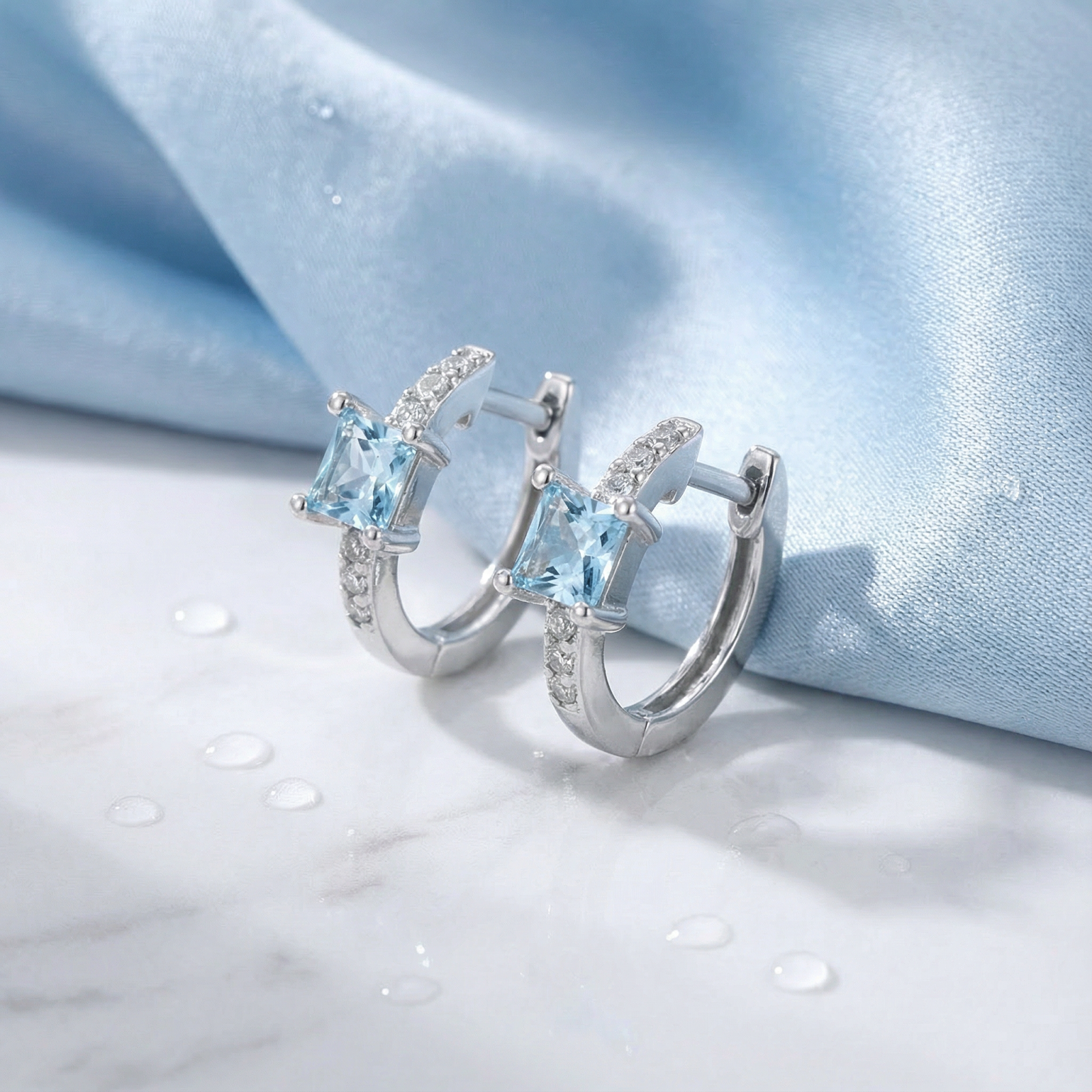 Side view of silver Aira huggie earrings with square-cut simulated aquamarine stones on a blue fabric background.