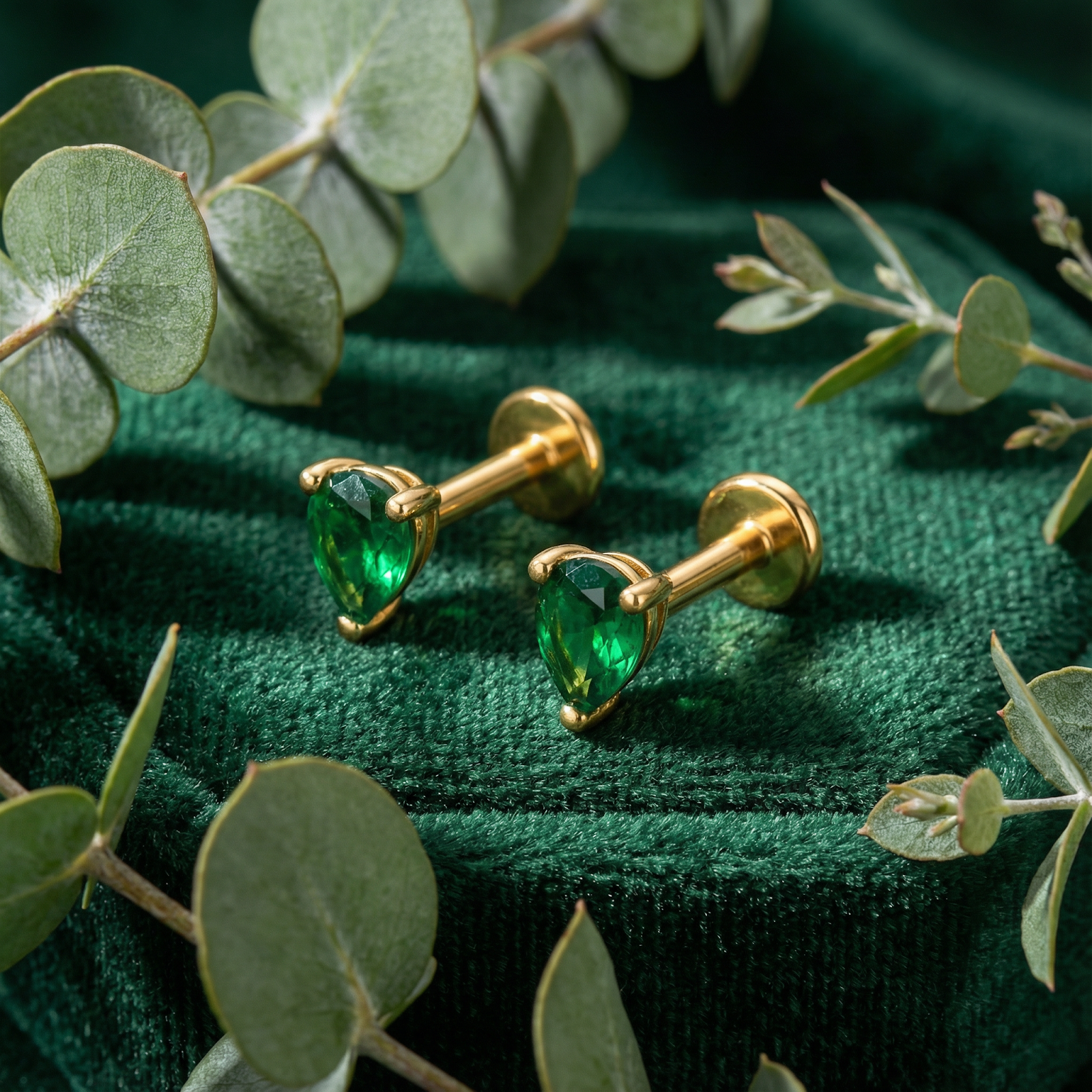 Pear-cut simulated emerald earrings in 18K gold vermeil on green velvet with eucalyptus leaves.