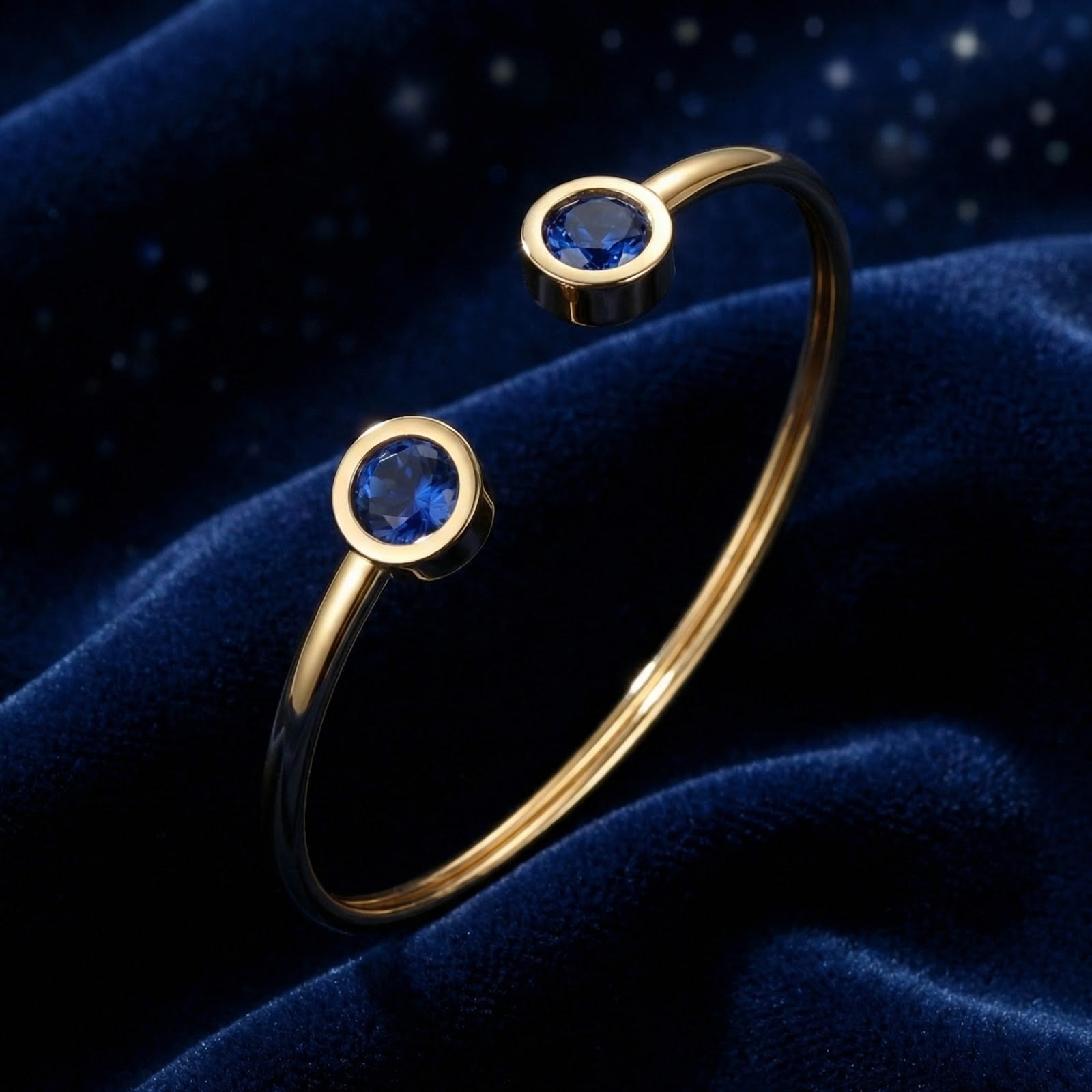 Gold stainless steel Verian bangle featuring two round blue simulated sapphires on blue velvet.
