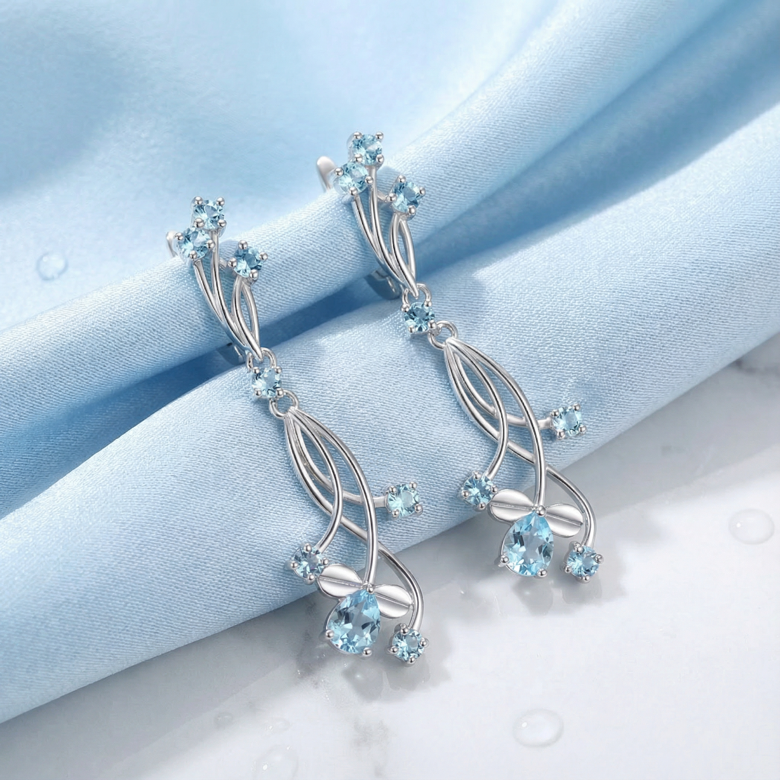 Lyriane drop earrings in S925 sterling silver with pear-cut simulated aquamarine on light blue silk fabric.