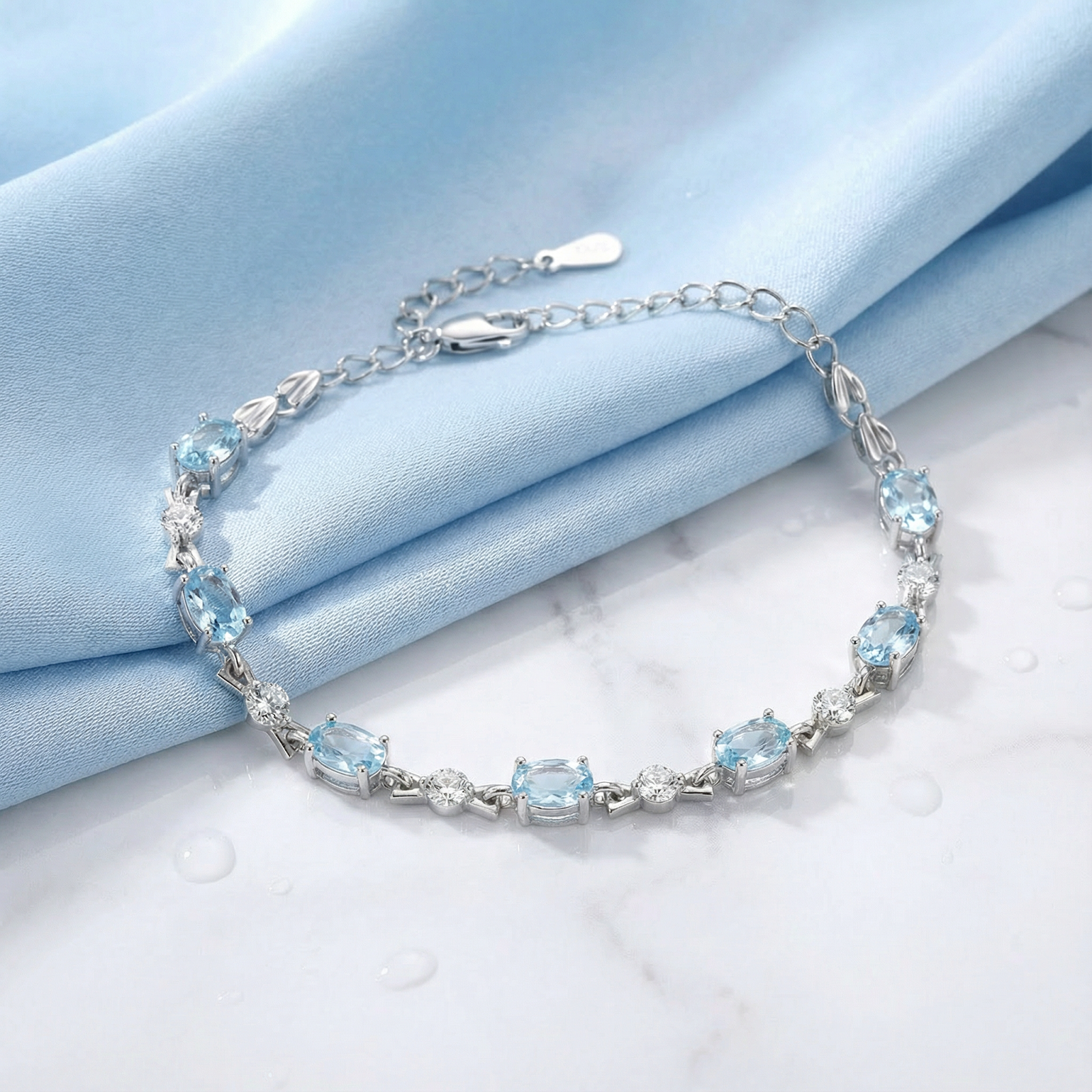 Sterling silver Melodia station bracelet with oval cut simulated aquamarine stones on a light blue fabric background.