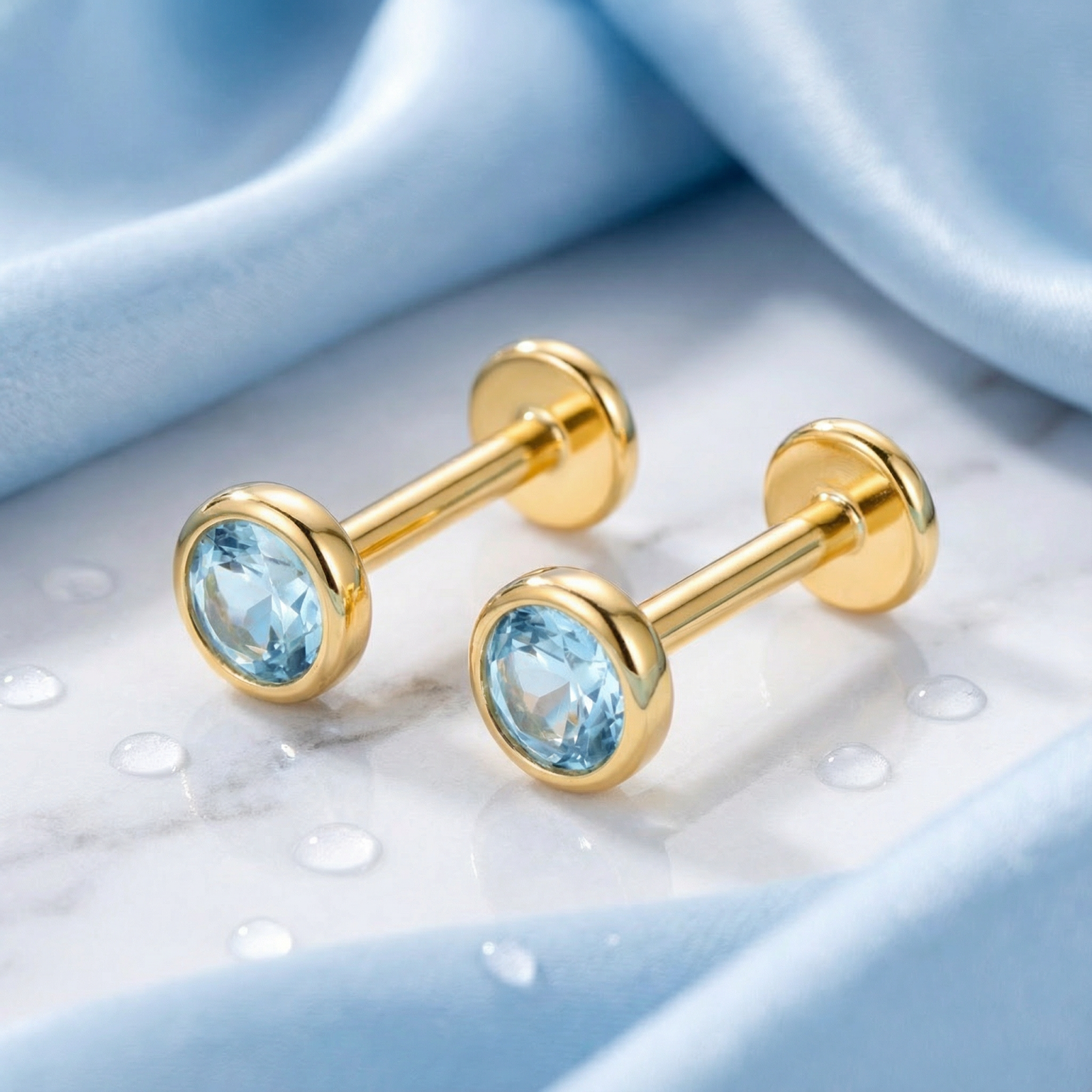 Side profile of round simulated aquamarine stud earrings in 18K gold vermeil on a blue fabric background.