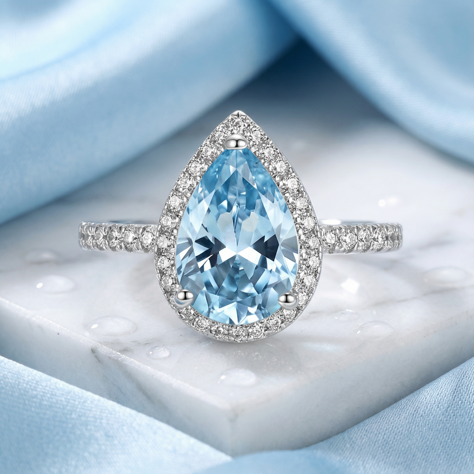Pear cut simulated aquamarine halo ring in S925 sterling silver on a marble and blue silk background.