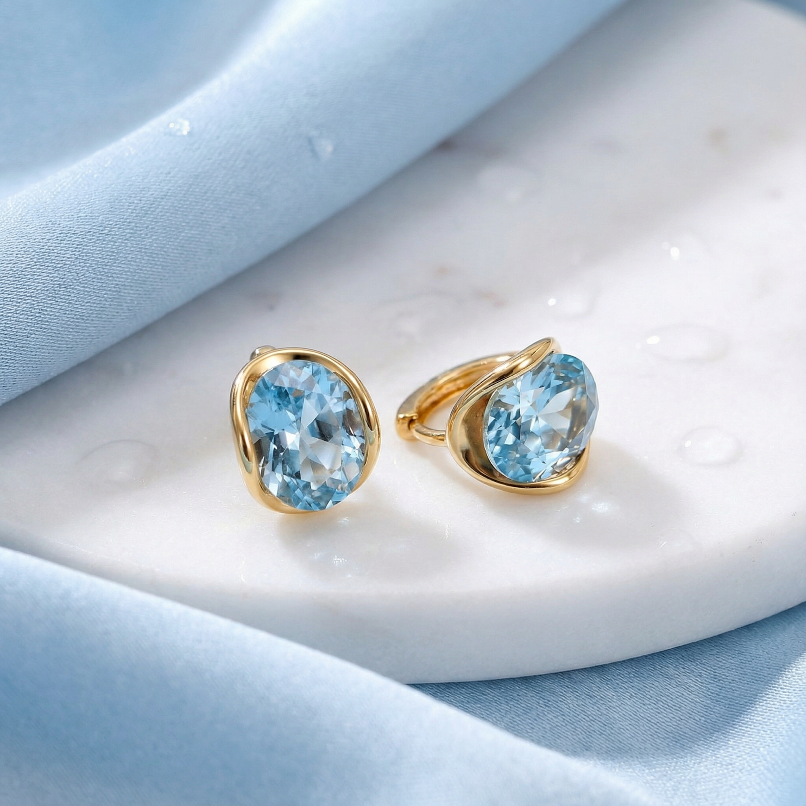 18K gold vermeil huggie earrings with oval simulated aquamarine stones on a marble and blue fabric background.