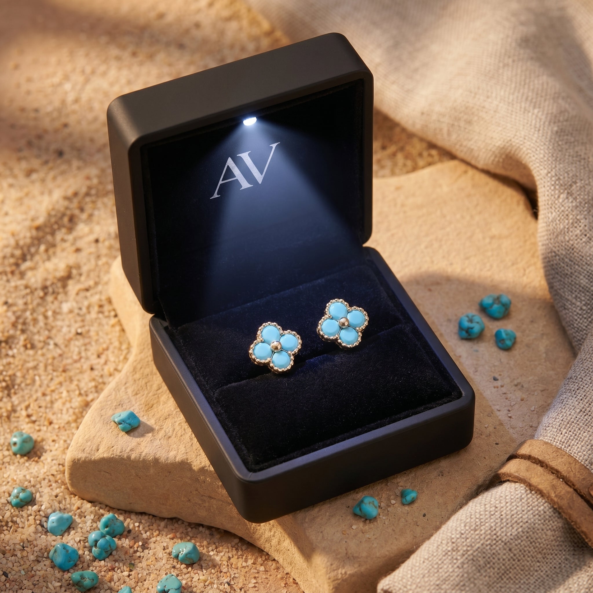 Gold turquoise clover stud earrings presented in a black light-up gift box on a sandy desert background.