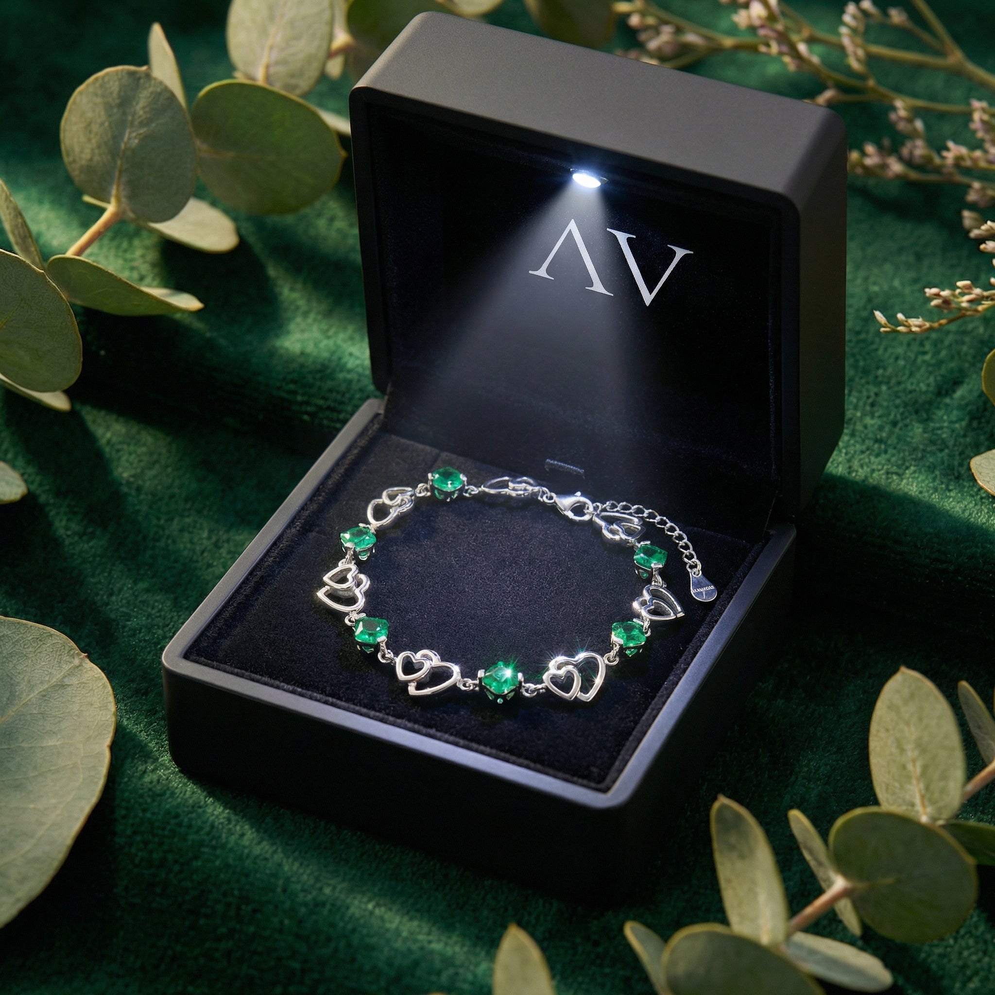 Silver heart bracelet with cushion cut simulated emeralds inside a black LED-lighted gift box on green velvet.
