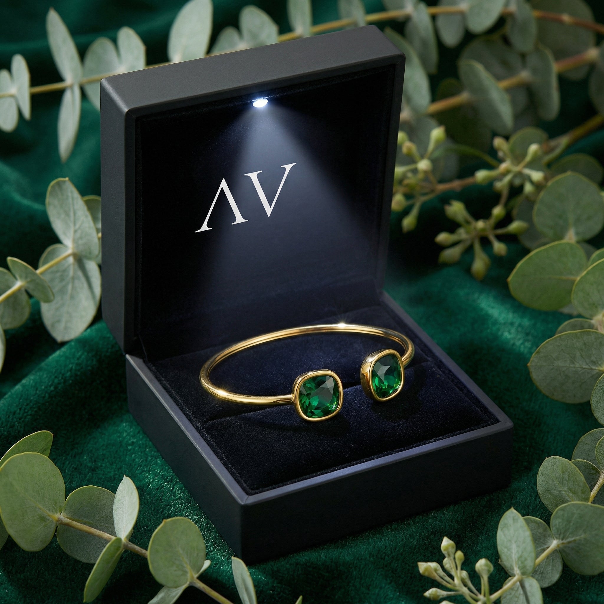Gold finish stainless steel bypass cuff bracelet with cushion cut simulated emeralds in a lighted black gift box.