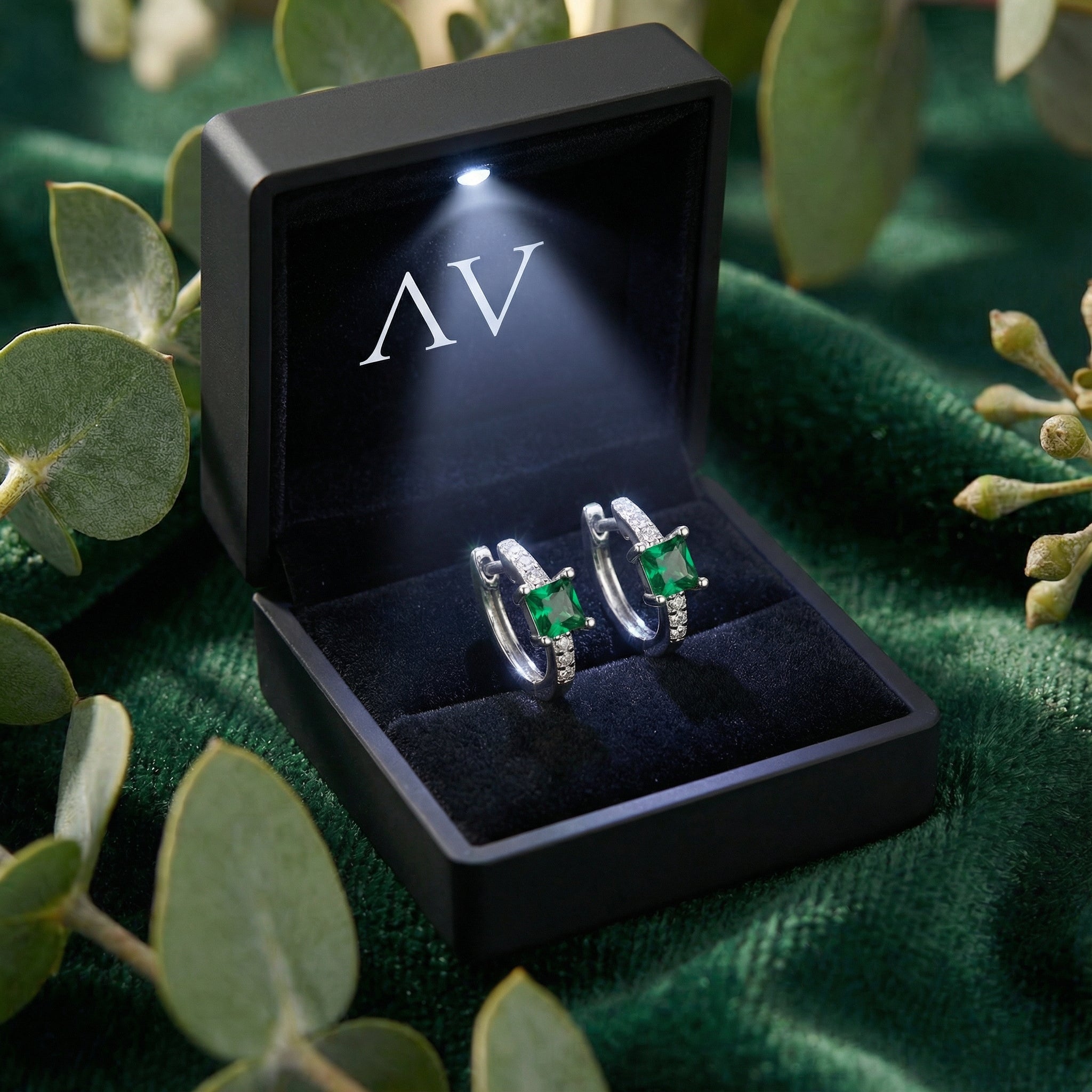 Sterling silver emerald huggie earrings inside a black LED-lit presentation box on green velvet.