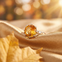 Round cut natural citrine ring in solid gold on gold satin fabric with an autumn leaf lifestyle background.