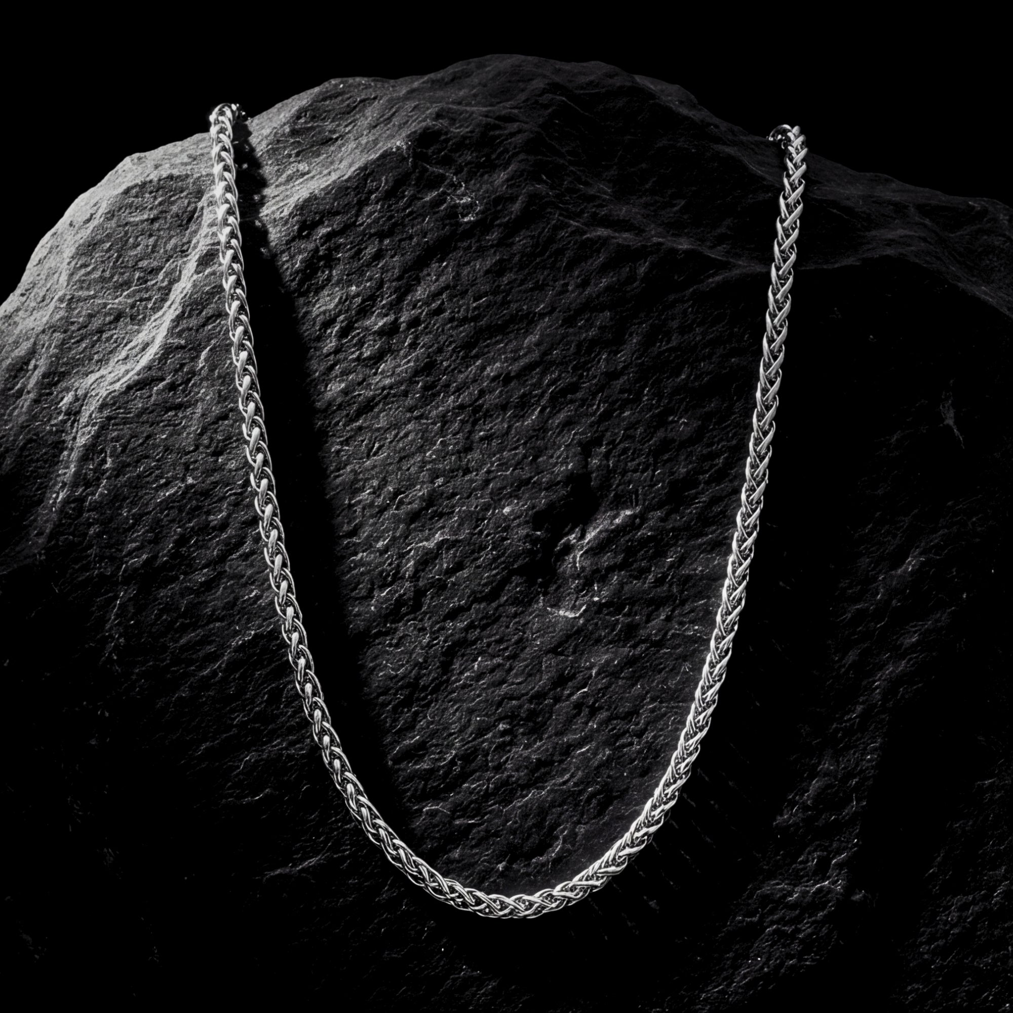 Silver stainless steel wheat weave chain necklace draped over a dark textured rock.