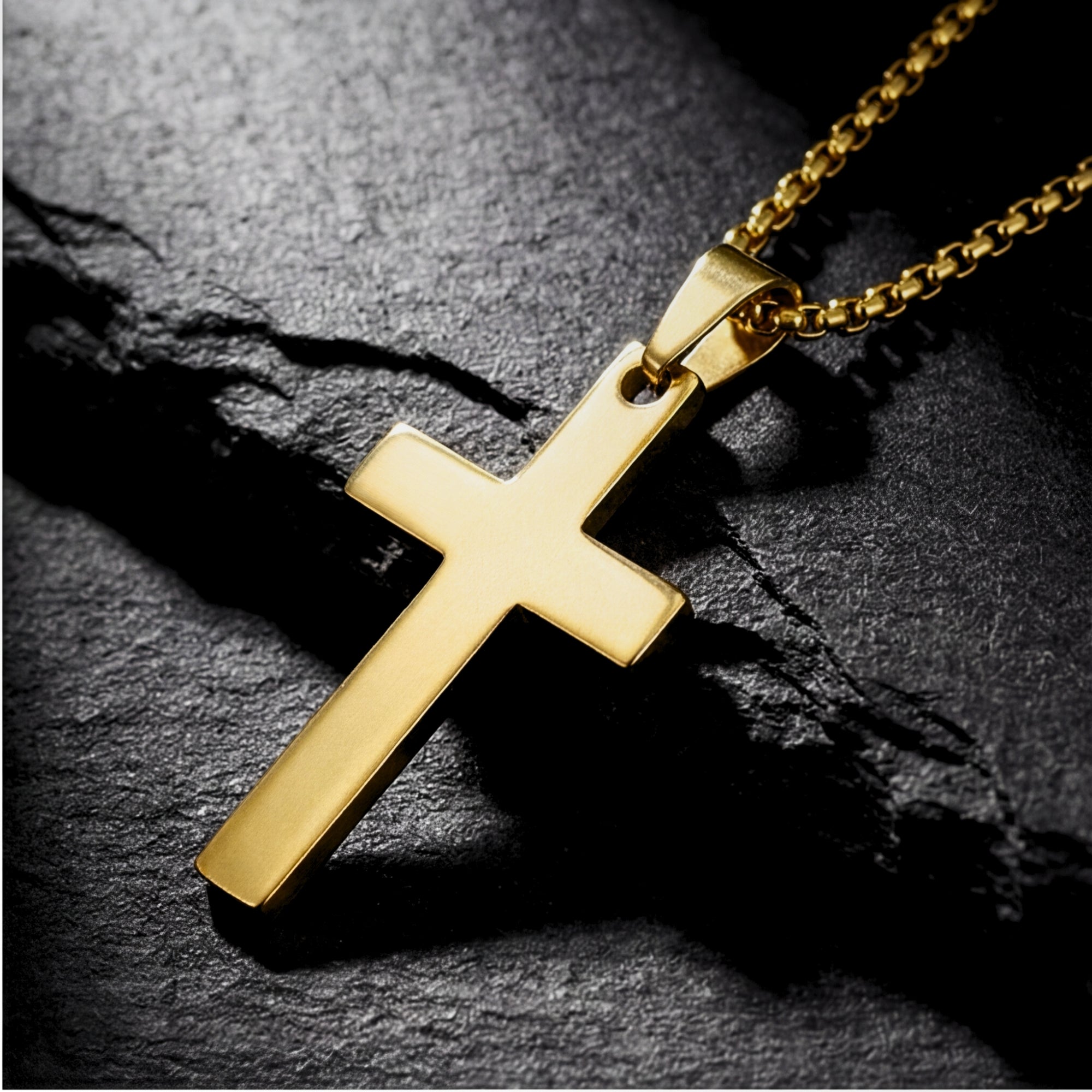 Gold stainless steel Apostle Cross pendant on a box chain, angled on a dark rugged stone surface.