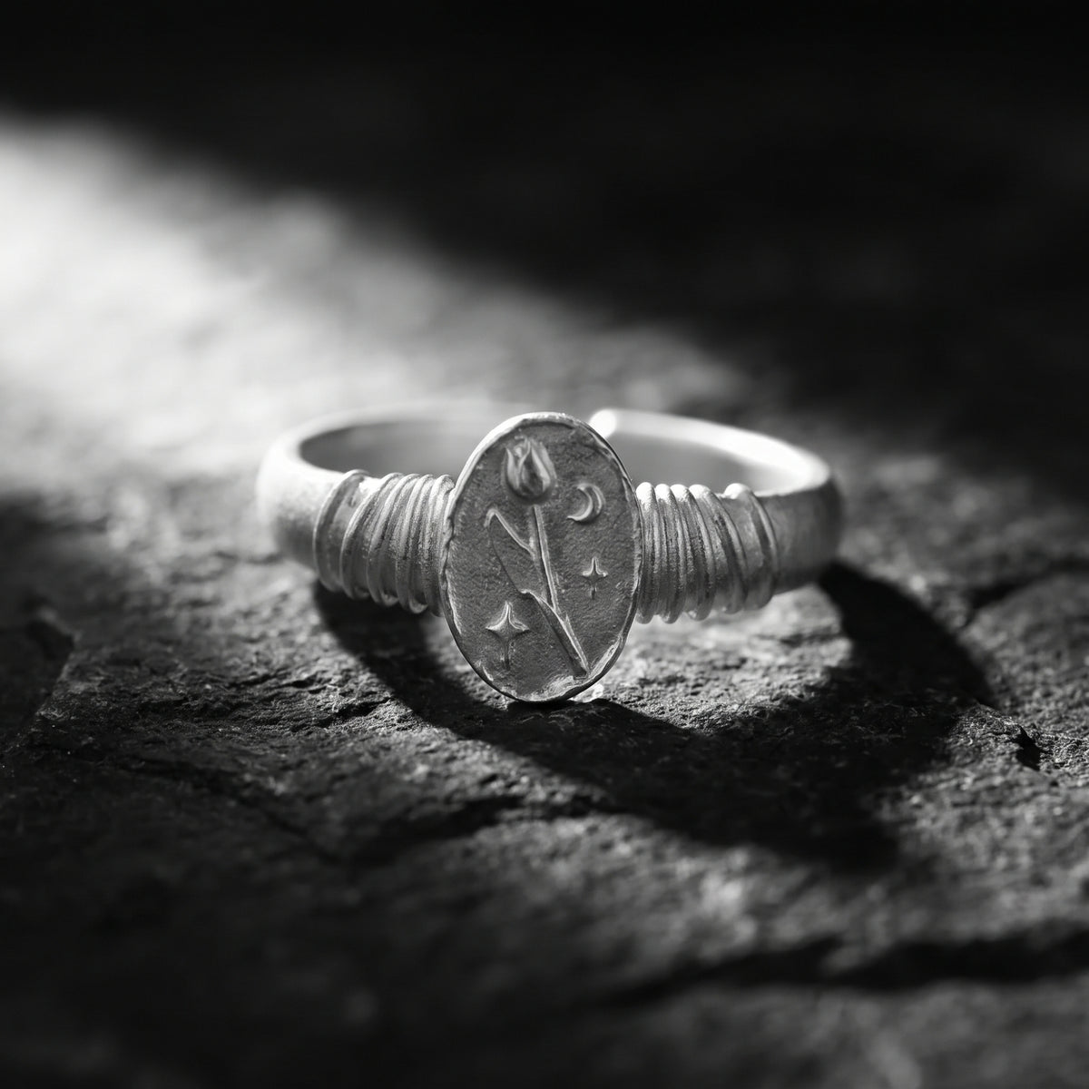 Silver signet ring on dark textured stone, showing an oval tulip, crescent moon, and starburst face.