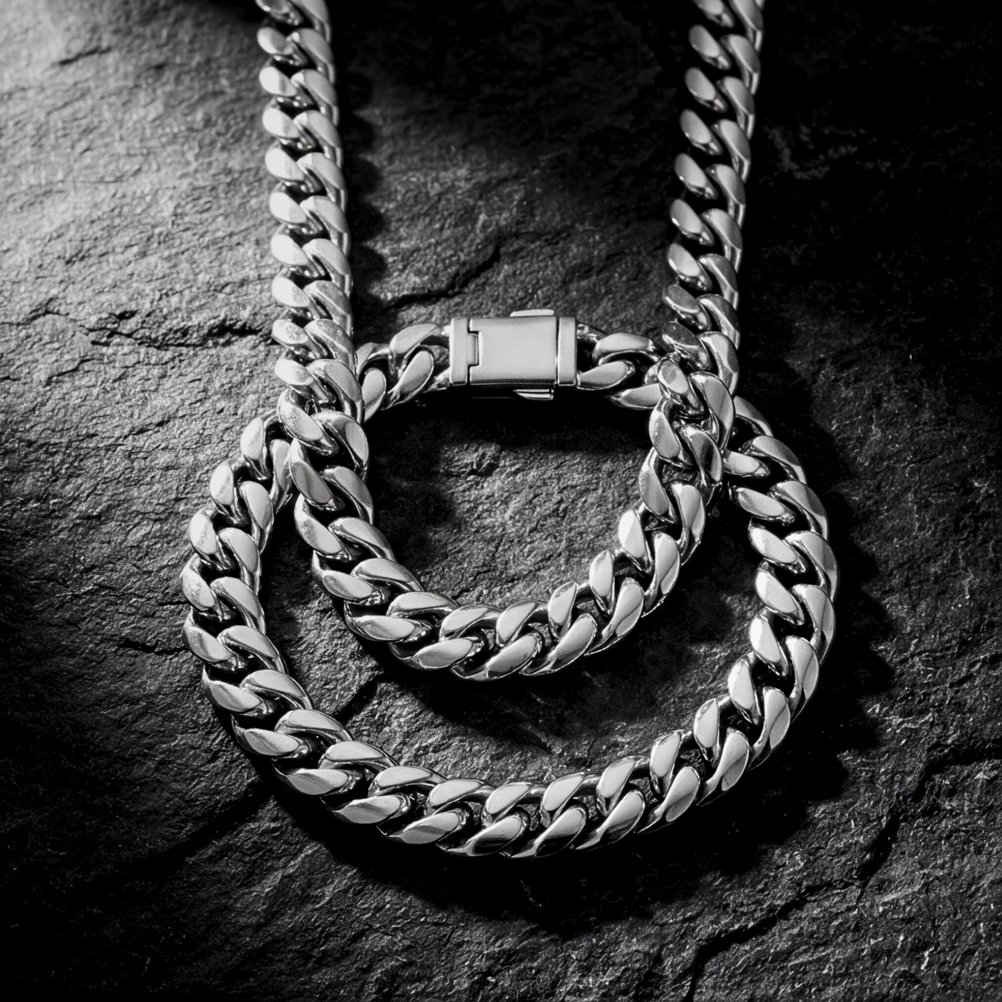 Polished silver 10mm stainless steel Cuban link chain and bracelet set coiled on a dark textured stone surface.