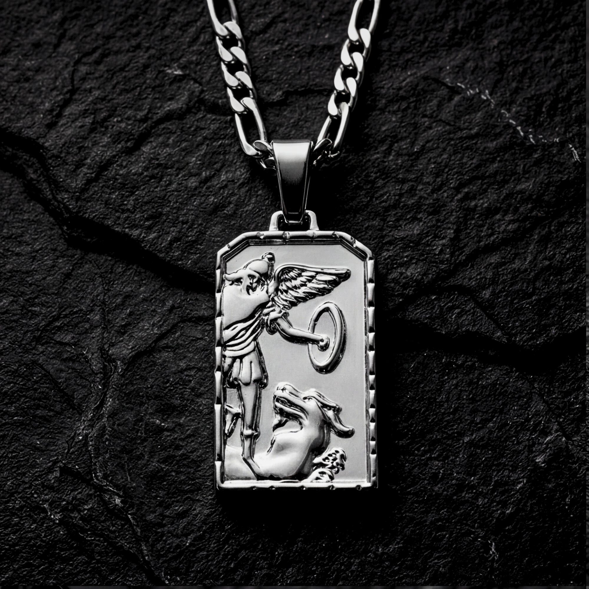 Silver stainless steel St. Michael's Victory pendant on a Figaro chain, resting on a dark textured stone background.