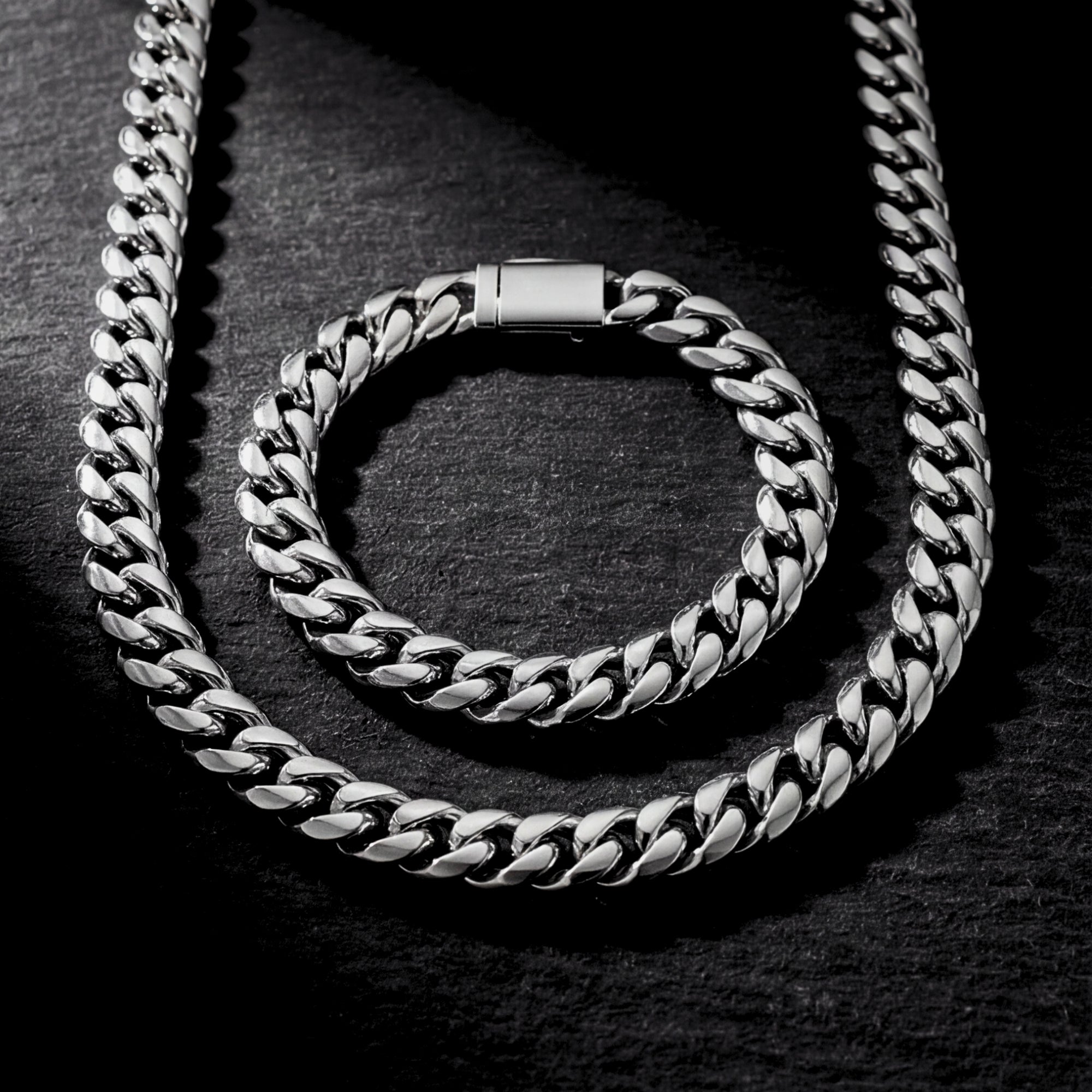 Silver stainless steel Cuban link chain and matching bracelet set displayed on a dark textured stone surface.