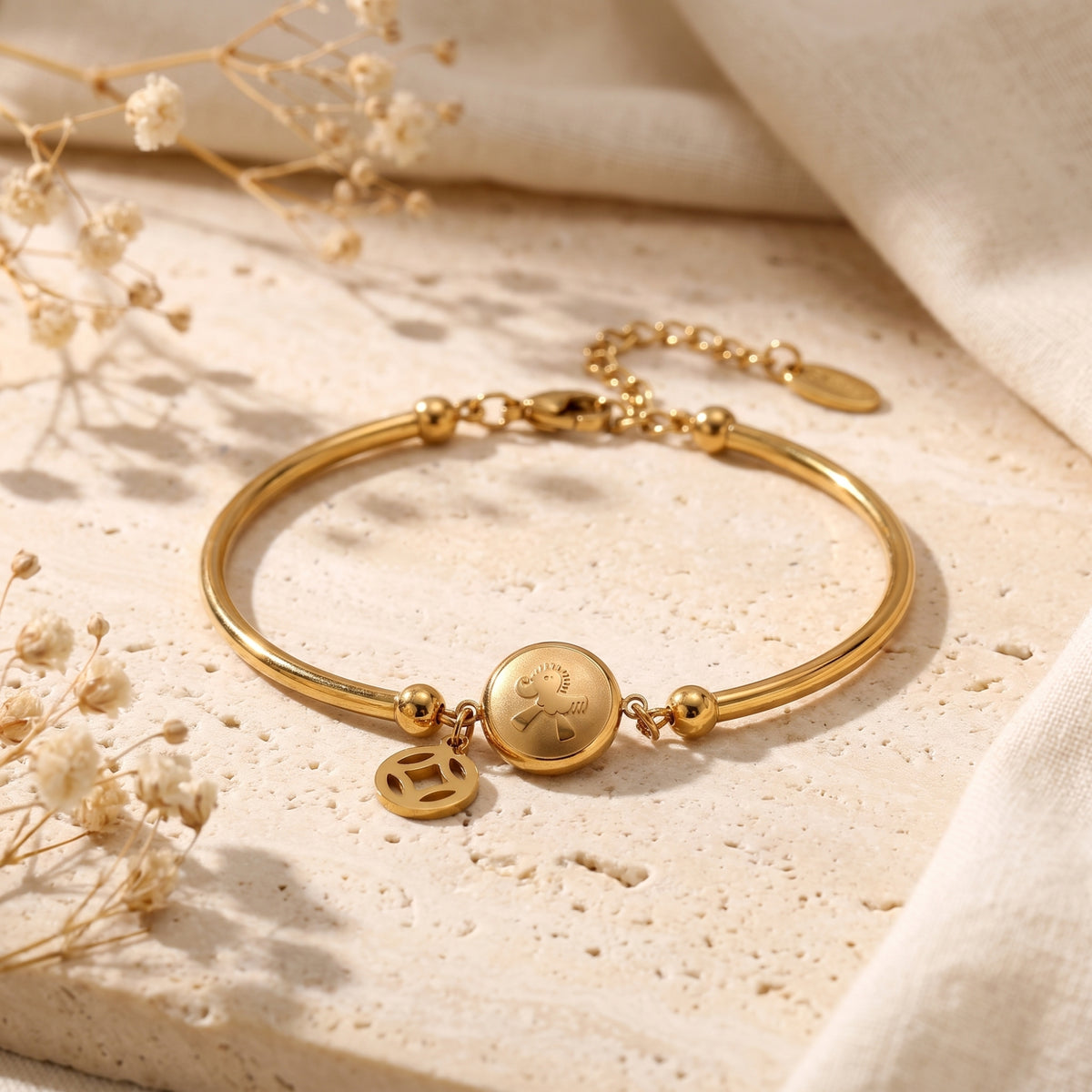 Gold-tone station bracelet with round medallion and openwork charm on a light textured stone surface with dried flowers.