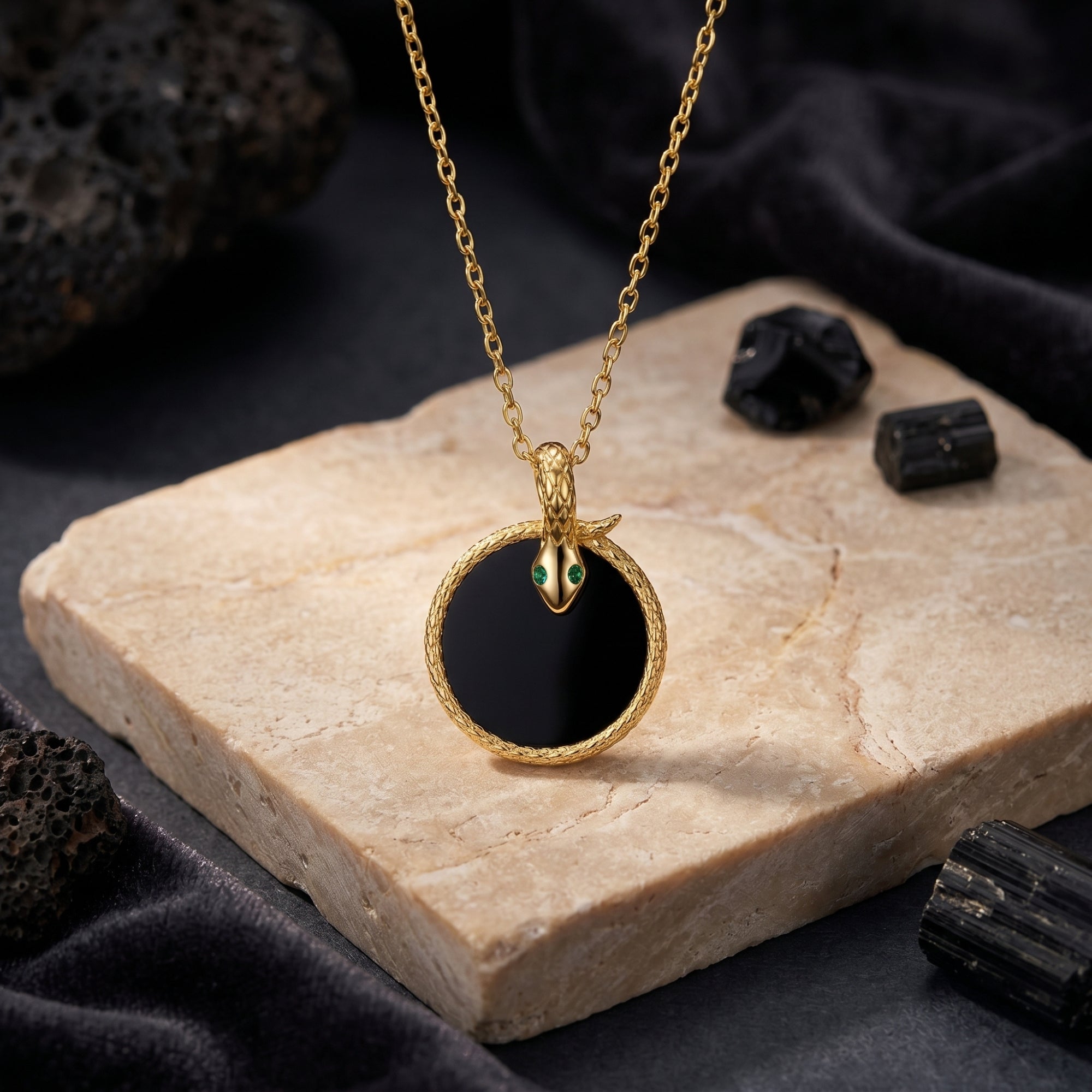Gold snake pendant with green eyes and round black onyx on a stone slab lifestyle background.