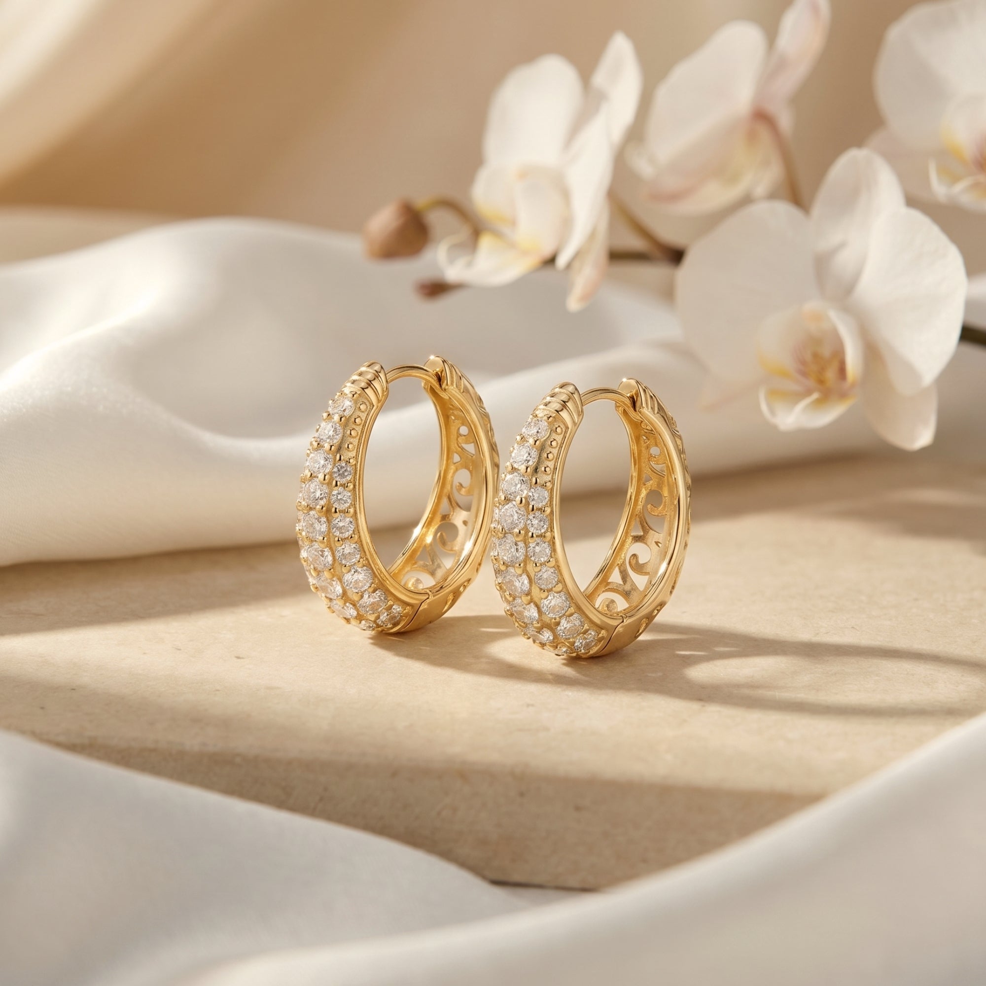 Gold round-cut moissanite hoop earrings on a stone surface with white orchids and silk.
