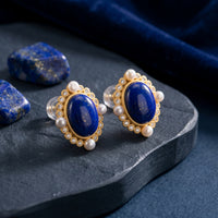 Oval natural lapis lazuli gold-plated stud earrings with pearls and crystals on a stone with raw lapis stones.