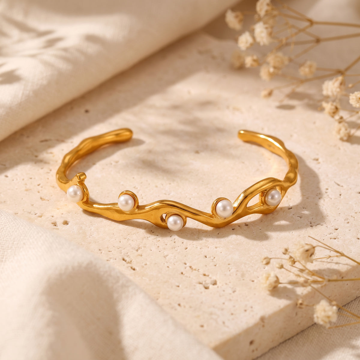 Gold-tone wave cuff bracelet with white simulated pearls resting on a textured stone display block.