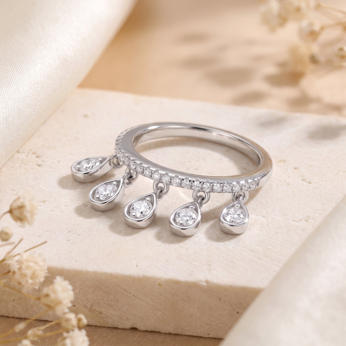 Silver pavé ring resting on a stone surface, with five dangling teardrop charms and round clear stones.