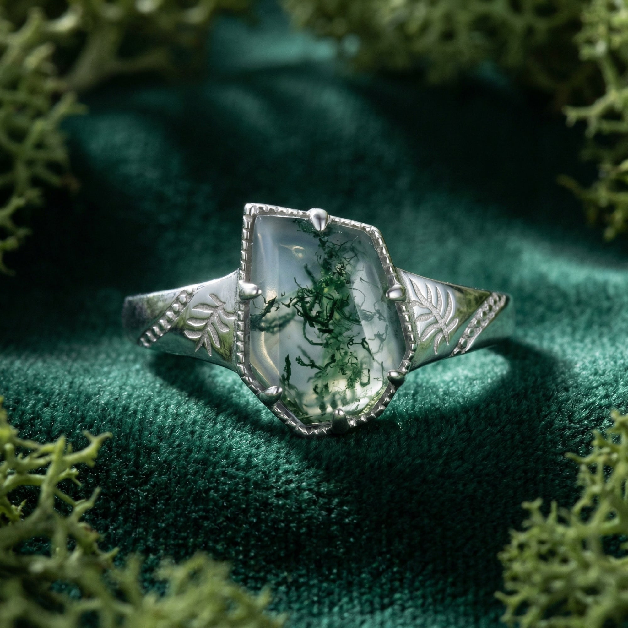 Shield cut moss agate ring in sterling silver with leaf engravings on a green velvet and moss background.