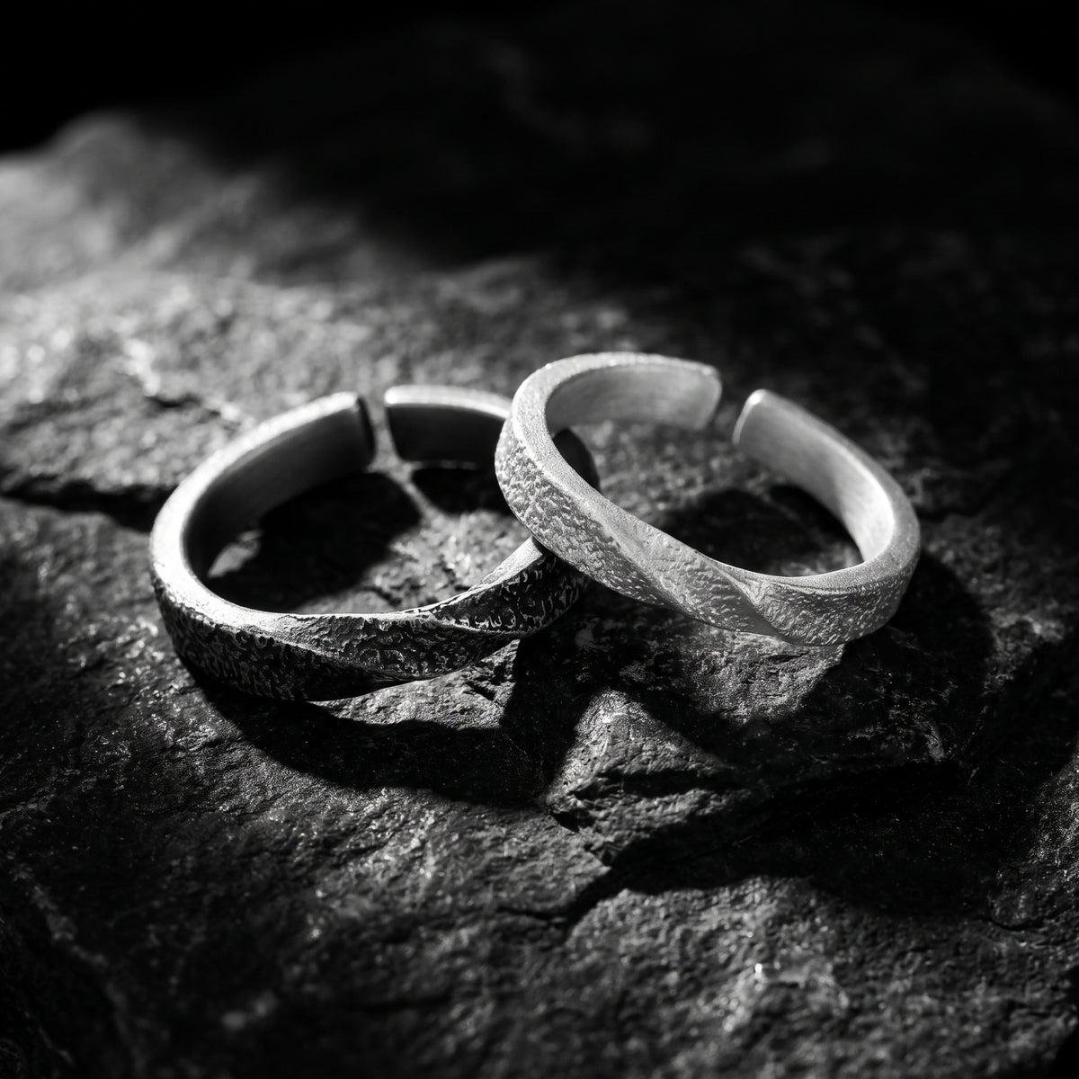 Two textured open-band rings, one dark-toned and one light-toned, crossing on rough rock.
