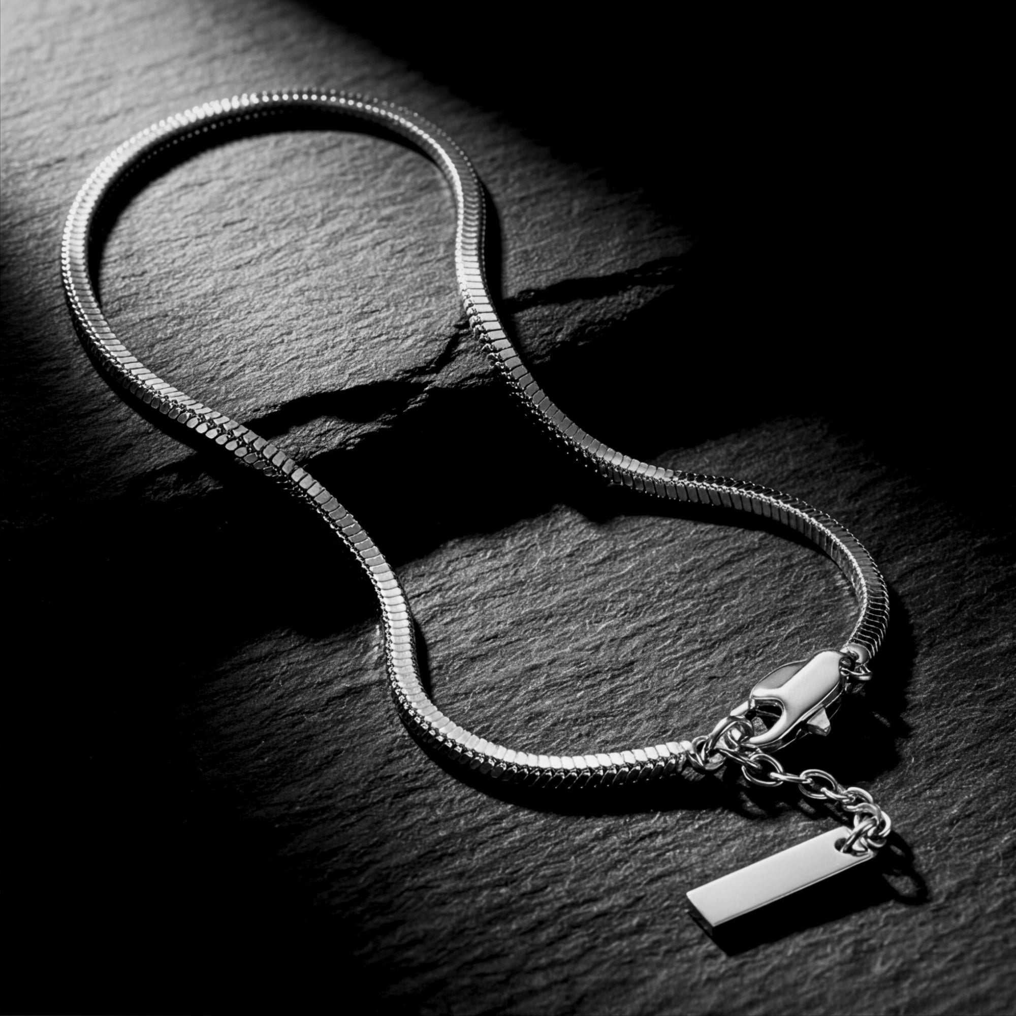 Silver stainless steel square link bracelet with lobster clasp on a dark textured stone surface.