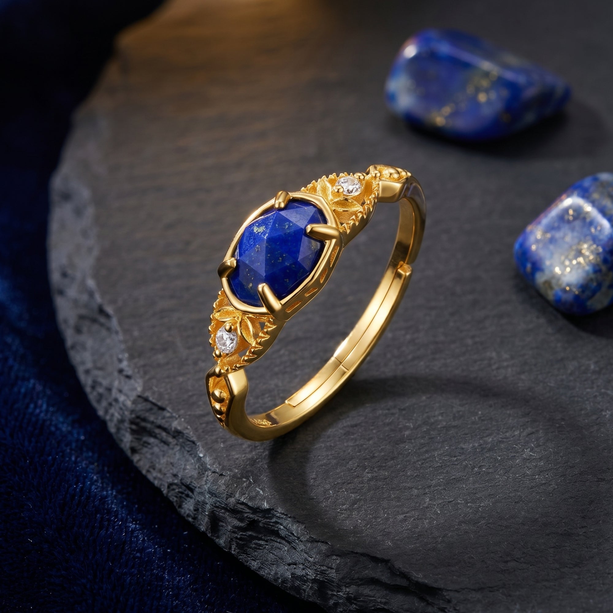 Gold Elarissa ring with a faceted oval lapis lazuli stone styled on a dark stone slab with loose blue gems.