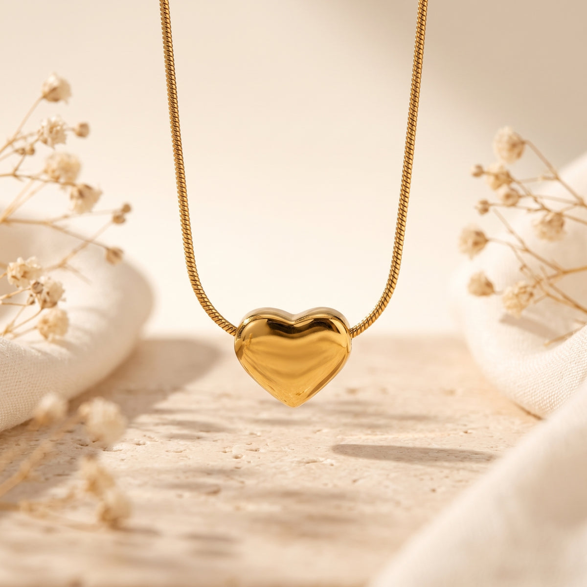 Close-up of a gold-tone puffy heart pendant on a snake chain, suspended above a stone surface with dried flowers.