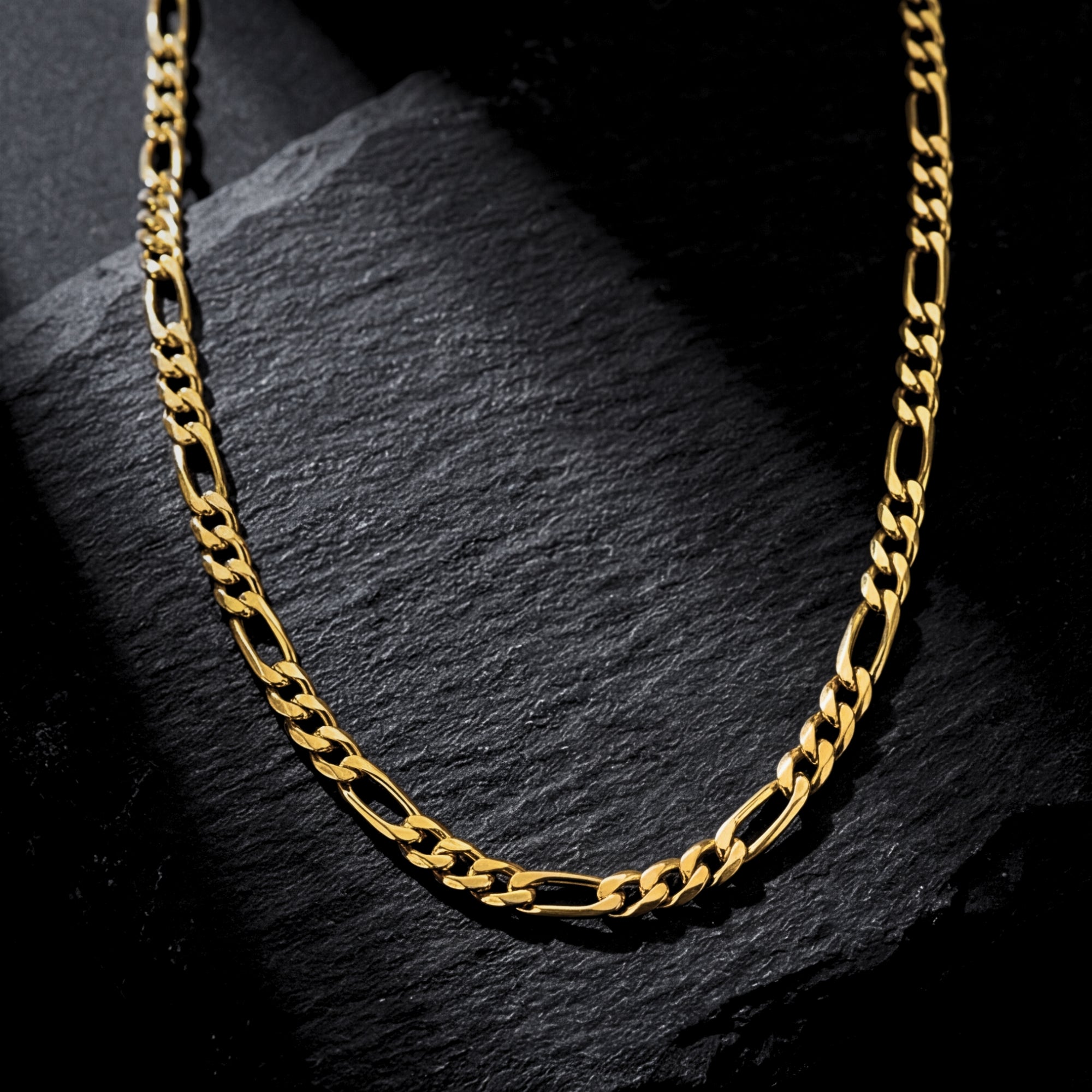 Gold 3mm Figaro chain in stainless steel displayed on a dark, textured stone background.
