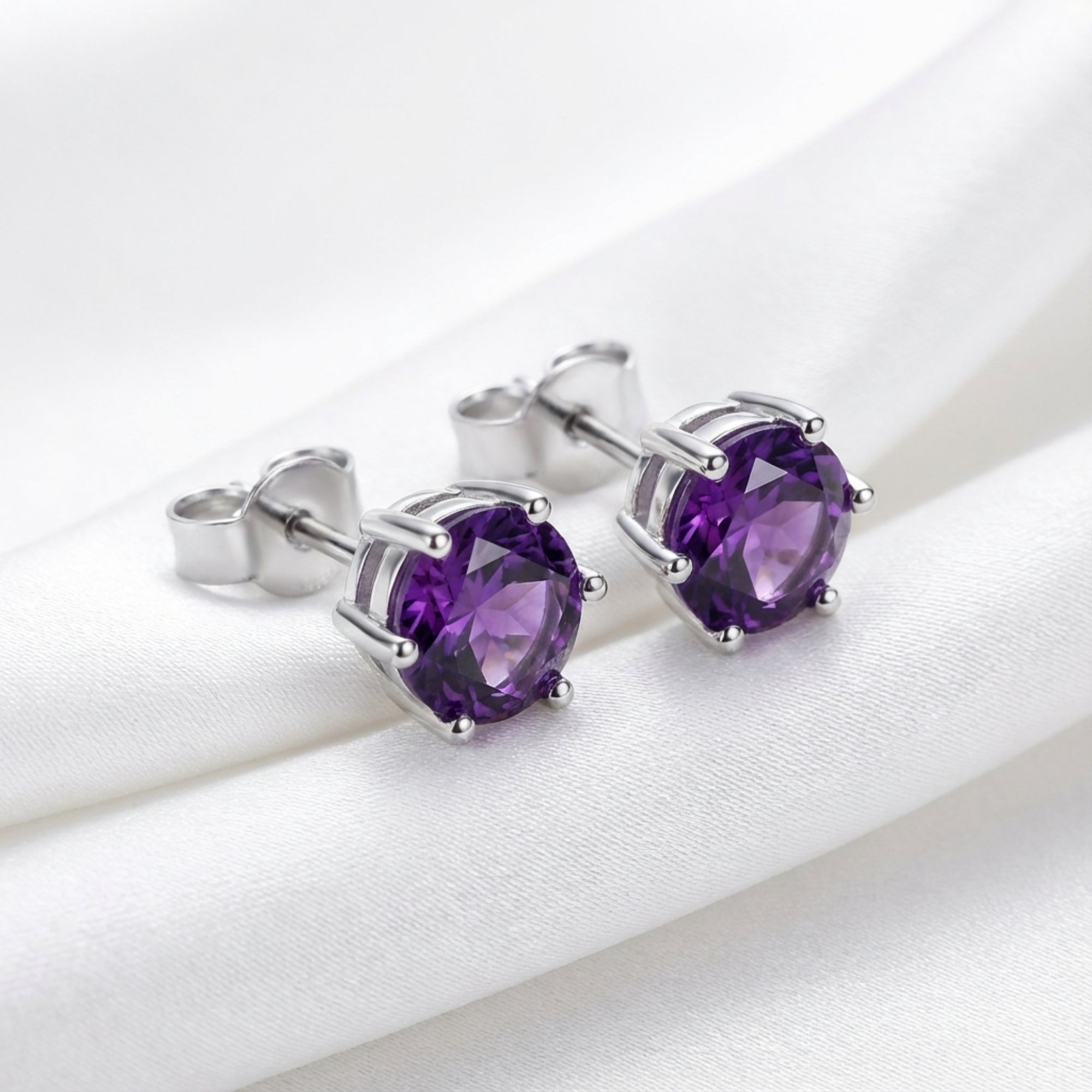 Pair of round cut simulated amethyst stud earrings in S925 sterling silver on white silk fabric.