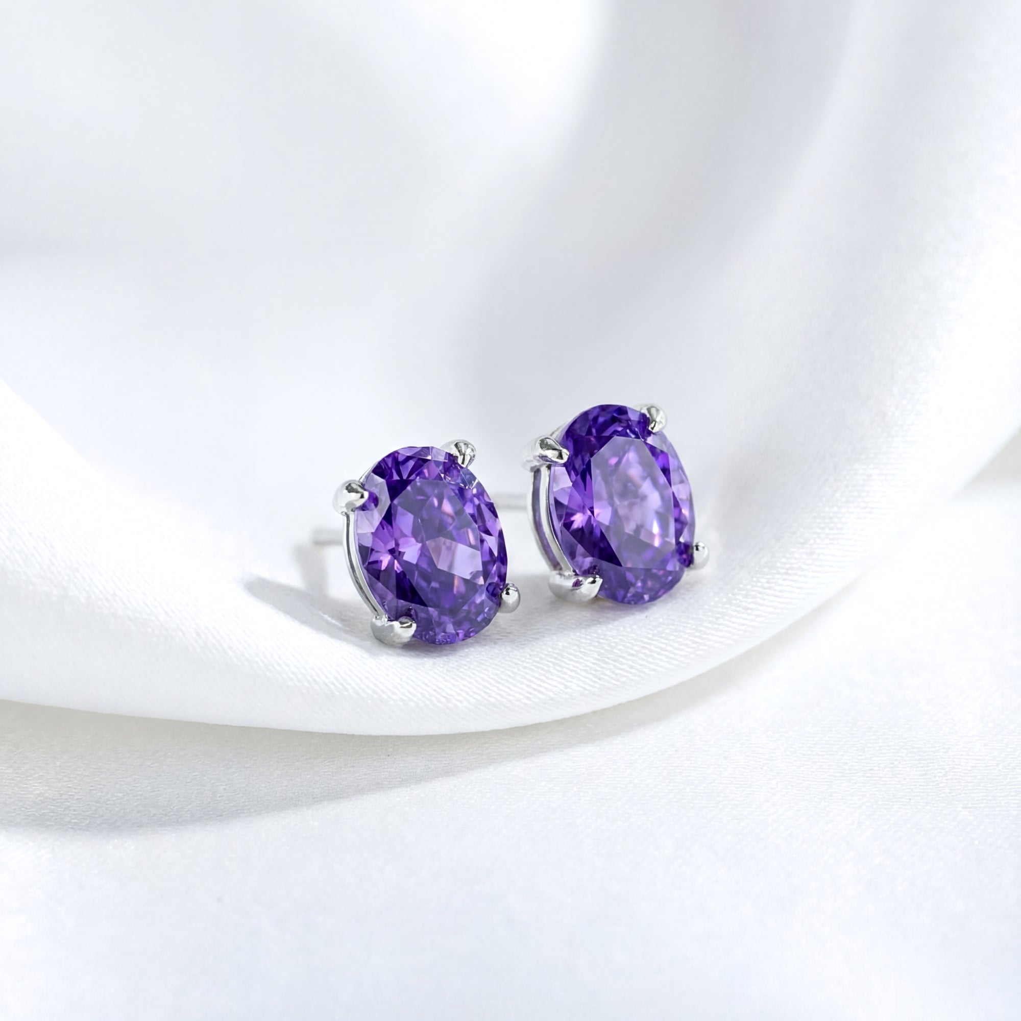 Oval cut purple simulated amethyst stud earrings in sterling silver resting on white silk fabric.
