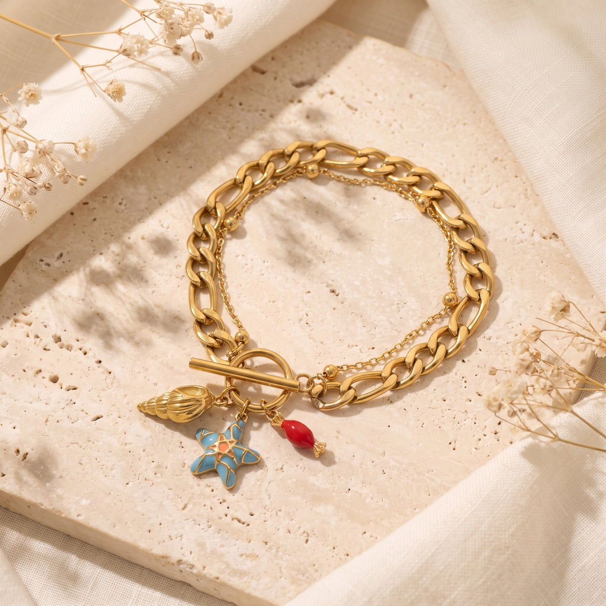 Gold-tone curb-chain charm bracelet with shell, blue starfish, and red charms on a textured stone slab.