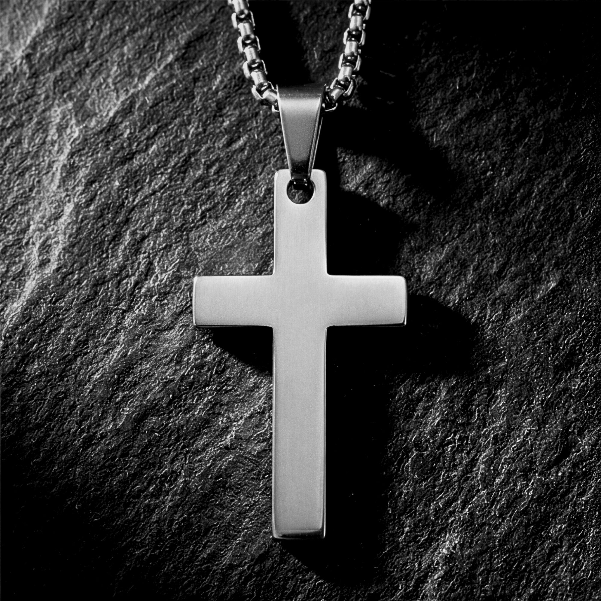 Silver stainless steel Apostle Cross pendant on a box chain, displayed on a dark textured stone surface.