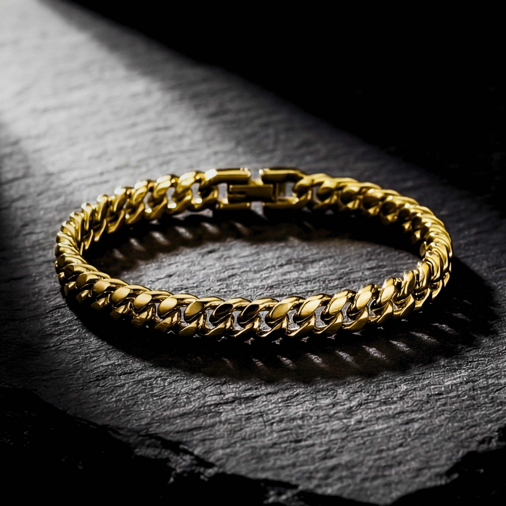 Gold stainless steel Cuban link bracelet resting on a dark textured stone surface with dramatic side lighting.