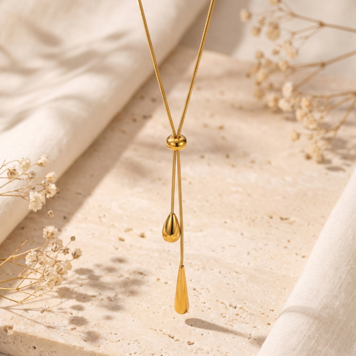 Gold-tone lariat necklace with a sliding bead and two cascading teardrop pendants displayed over textured stone.