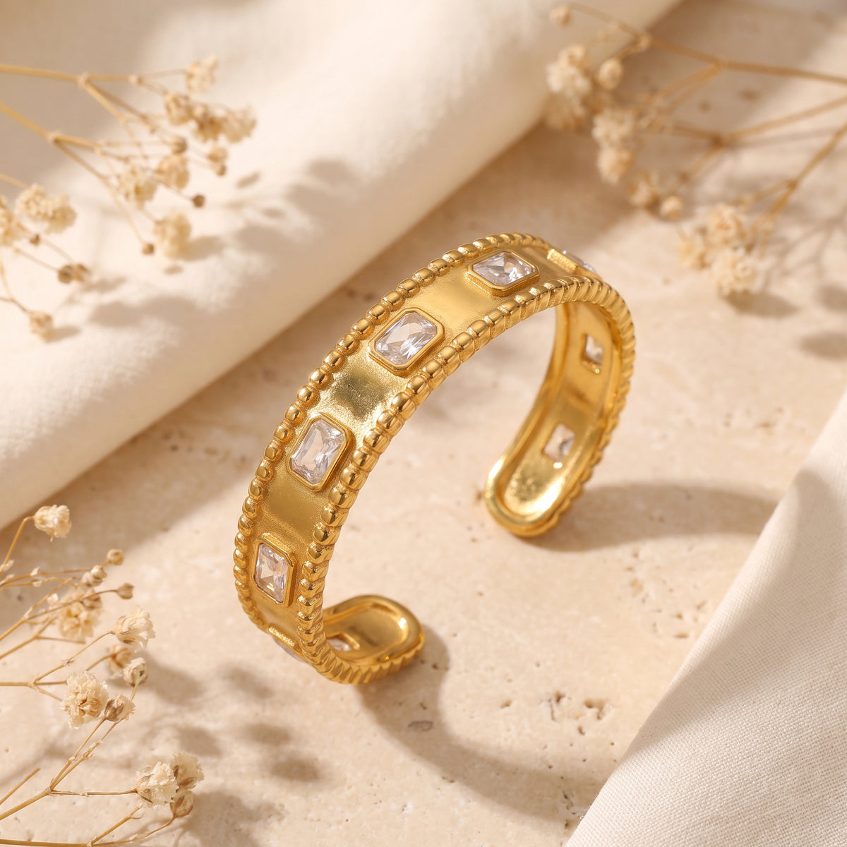 Angled close-up of a gold-tone cuff bracelet with beaded edges and bezel-set rectangular clear stones.