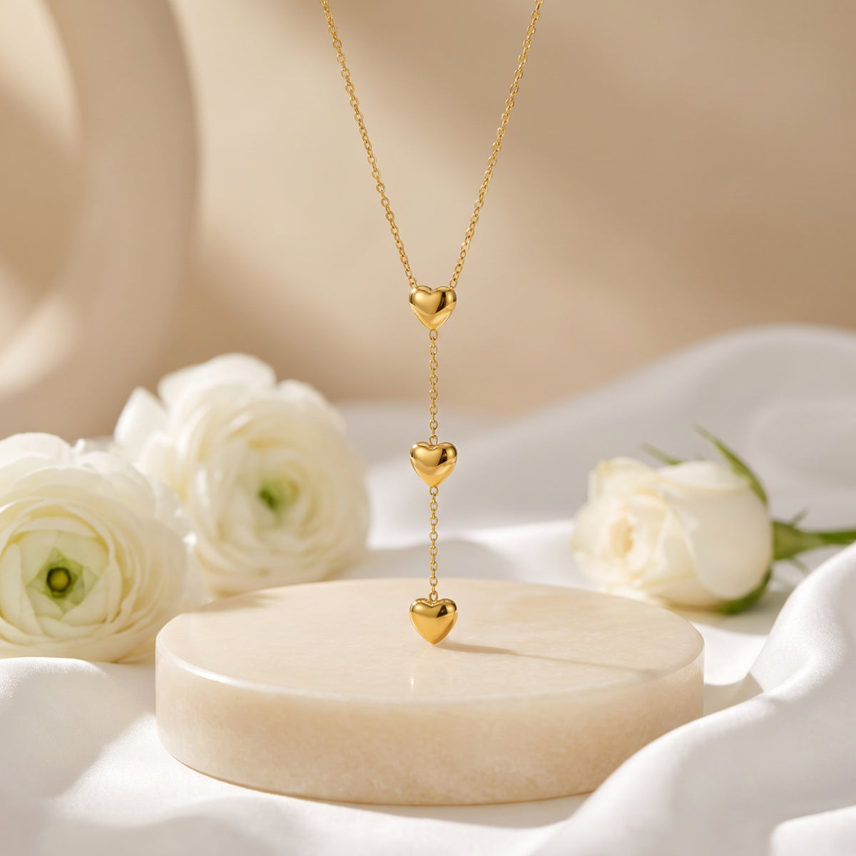 Gold-tone triple heart lariat necklace hanging above a round pedestal with white flowers