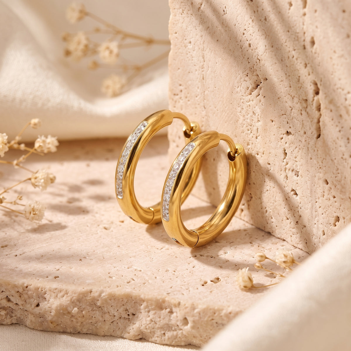 Pair of gold-tone pavé huggie hoop earrings standing against a beige textured stone block.