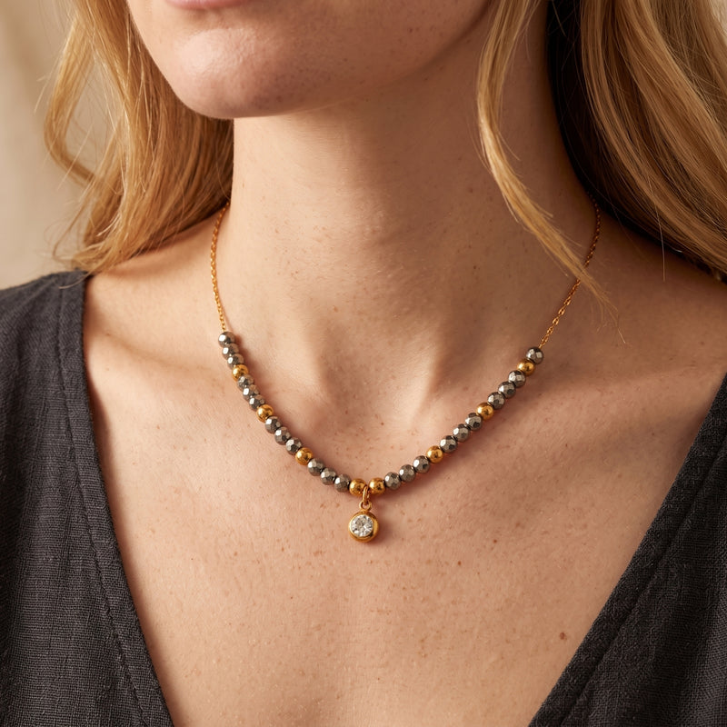 Model wearing a gold-tone beaded necklace with faceted gray beads and a round pendant over a dark top.