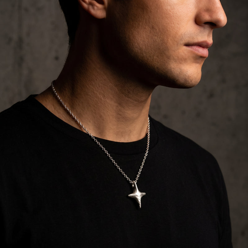 Man in side profile wearing a silver four-point starburst pendant on a chain over a black shirt.