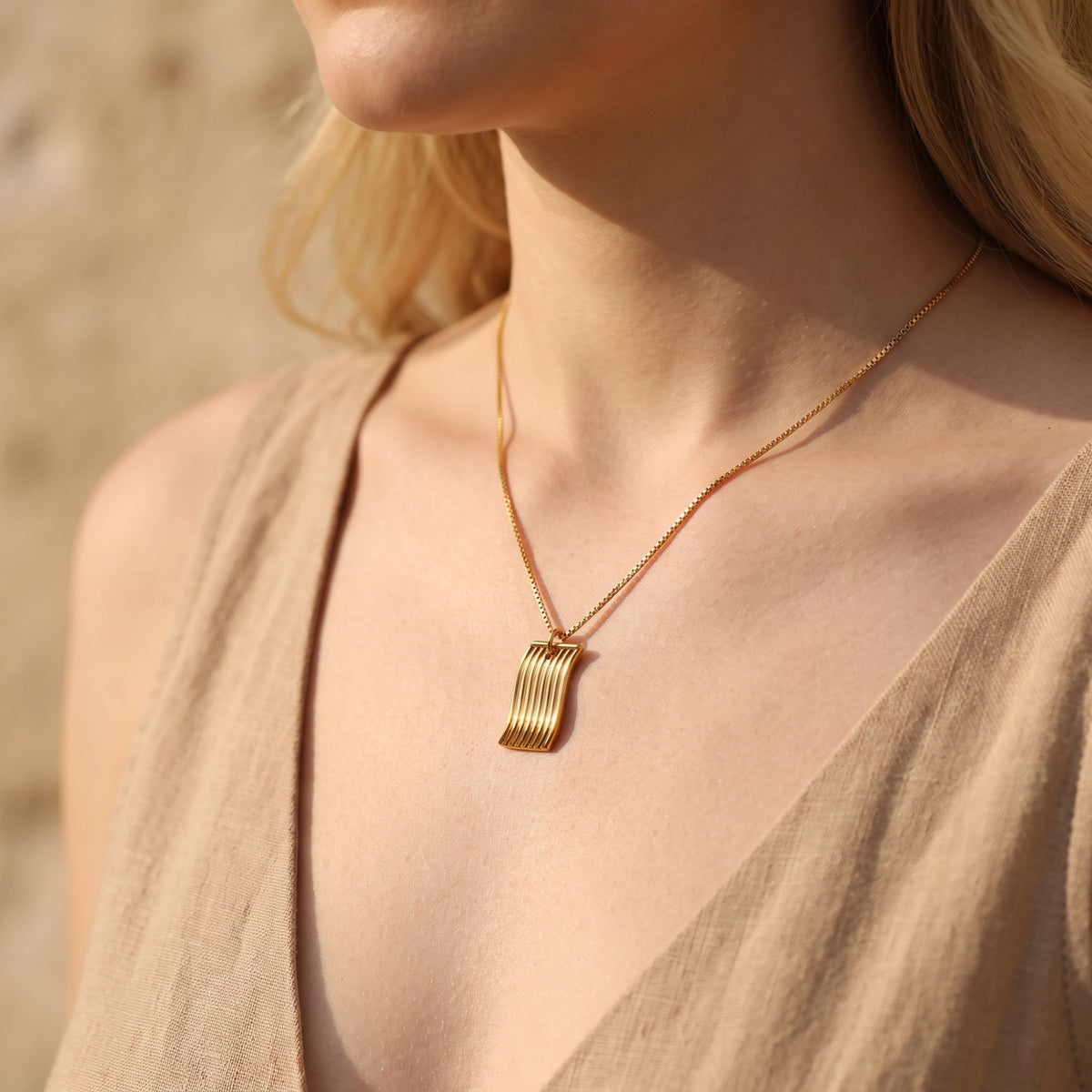 Gold-tone fluid wave pendant necklace on a box chain worn on a woman's neckline.